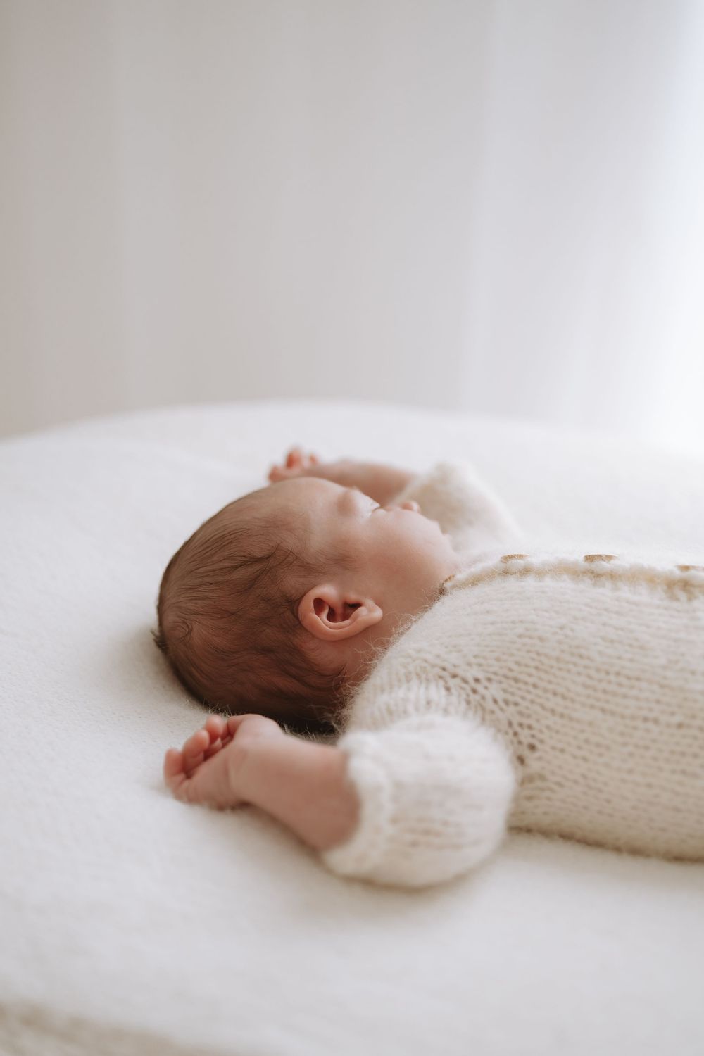 A sleeping newborn baby wrapped in a white knit blanket lies on a soft white surface in a minimalist studio setting.