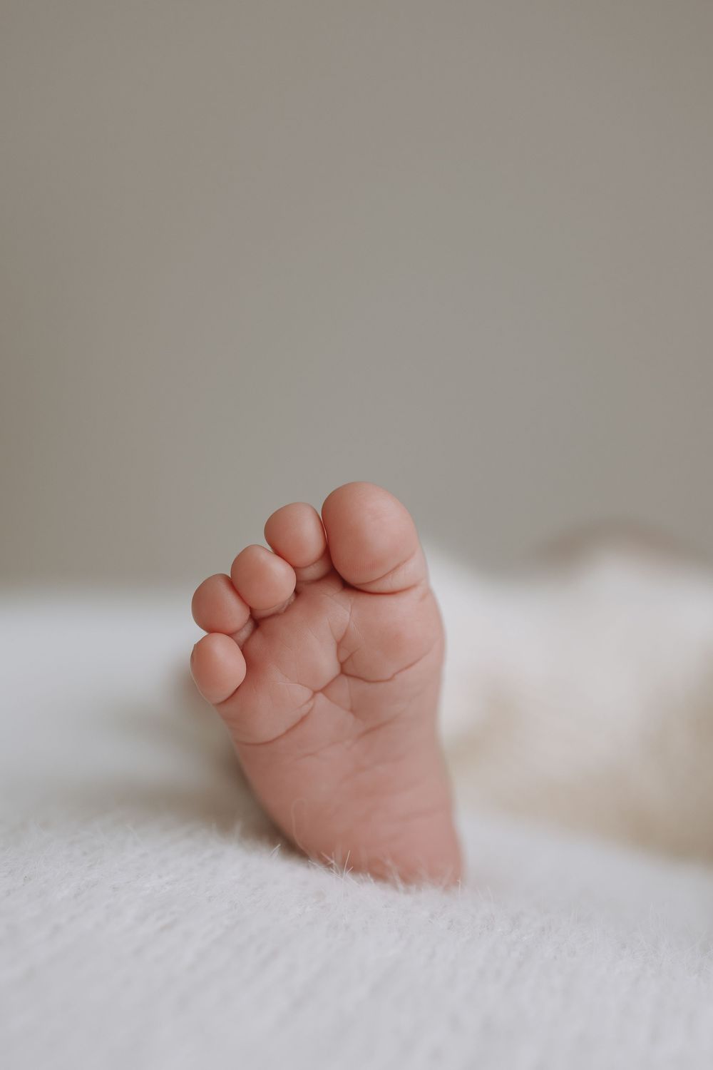 Tiny baby foot with delicate toes resting on soft white fur blanket against neutral background.