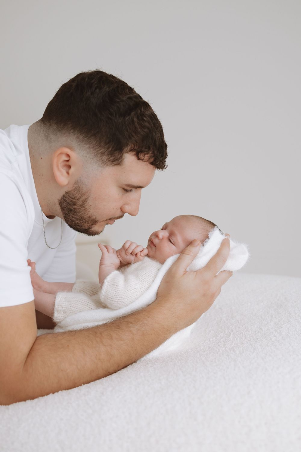 Parent in white shirt tenderly holding and gazing at newborn baby wrapped in soft blanket against light background.