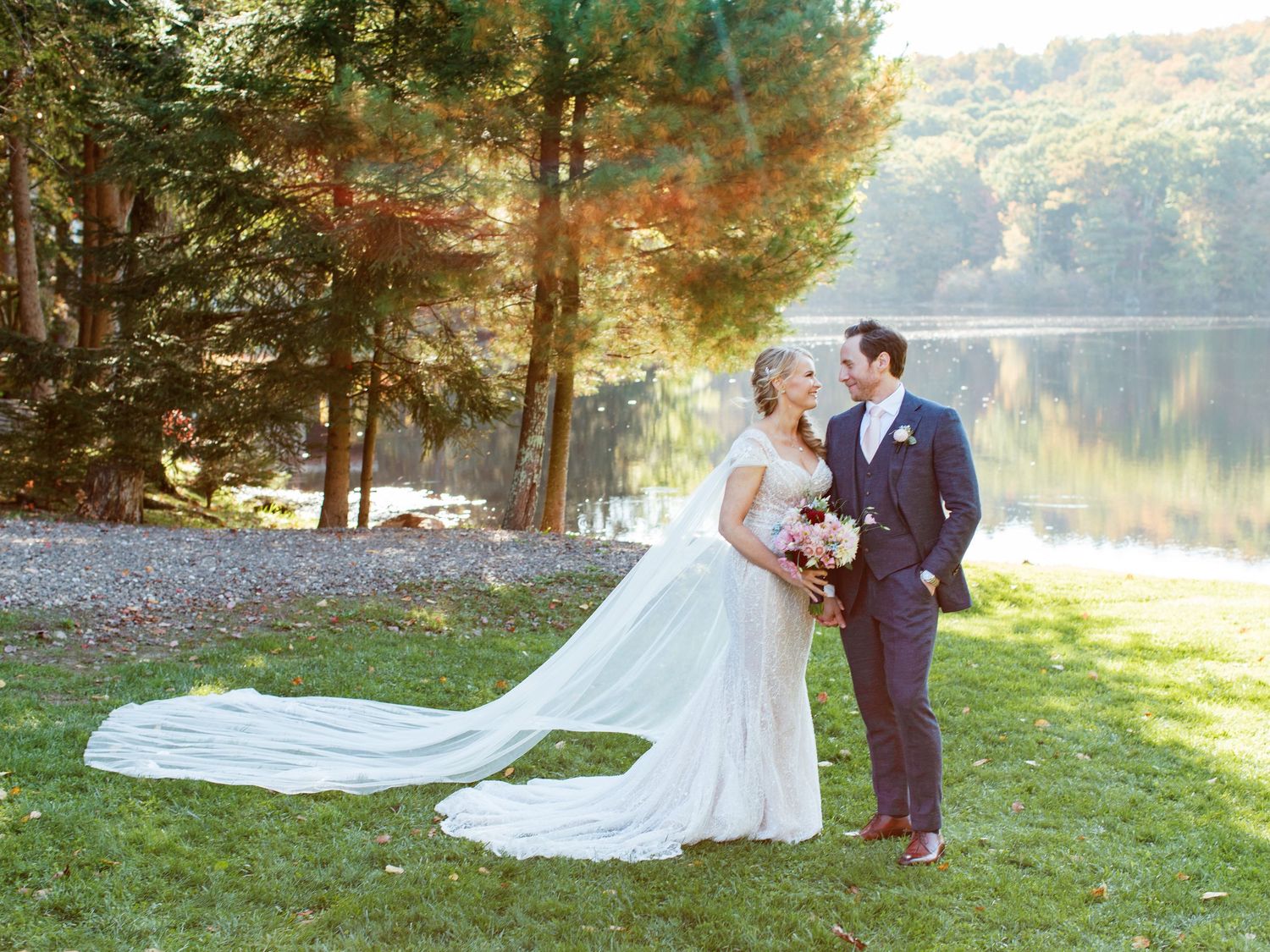 A romantic lakeside wedding photo with a long flowing dress train against a backdrop of pine trees and water.