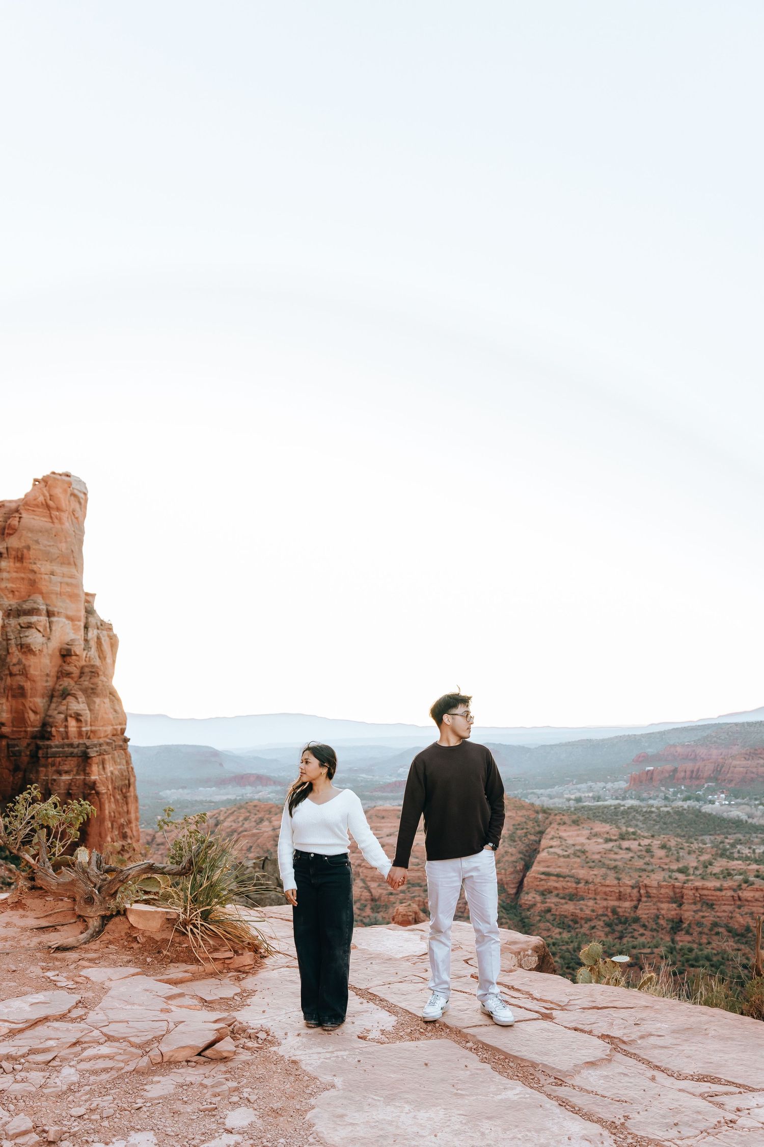 Two people walk hand in hand along a scenic desert cliff edge at sunset.