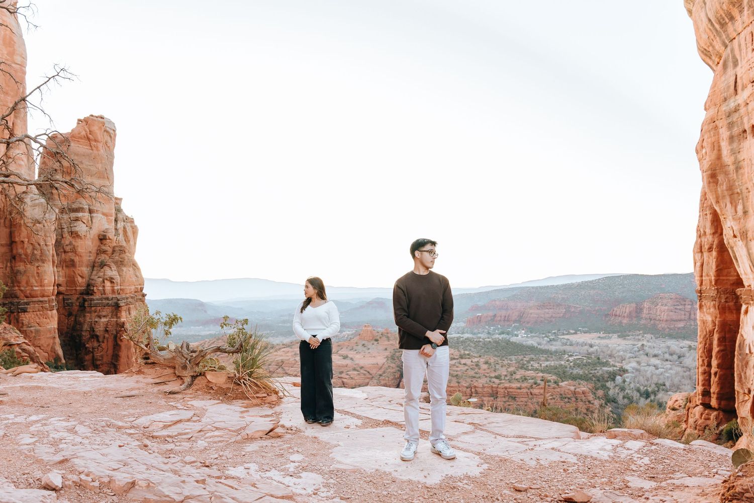 Two people enjoy panoramic views from a high vantage point among towering red rock formations.