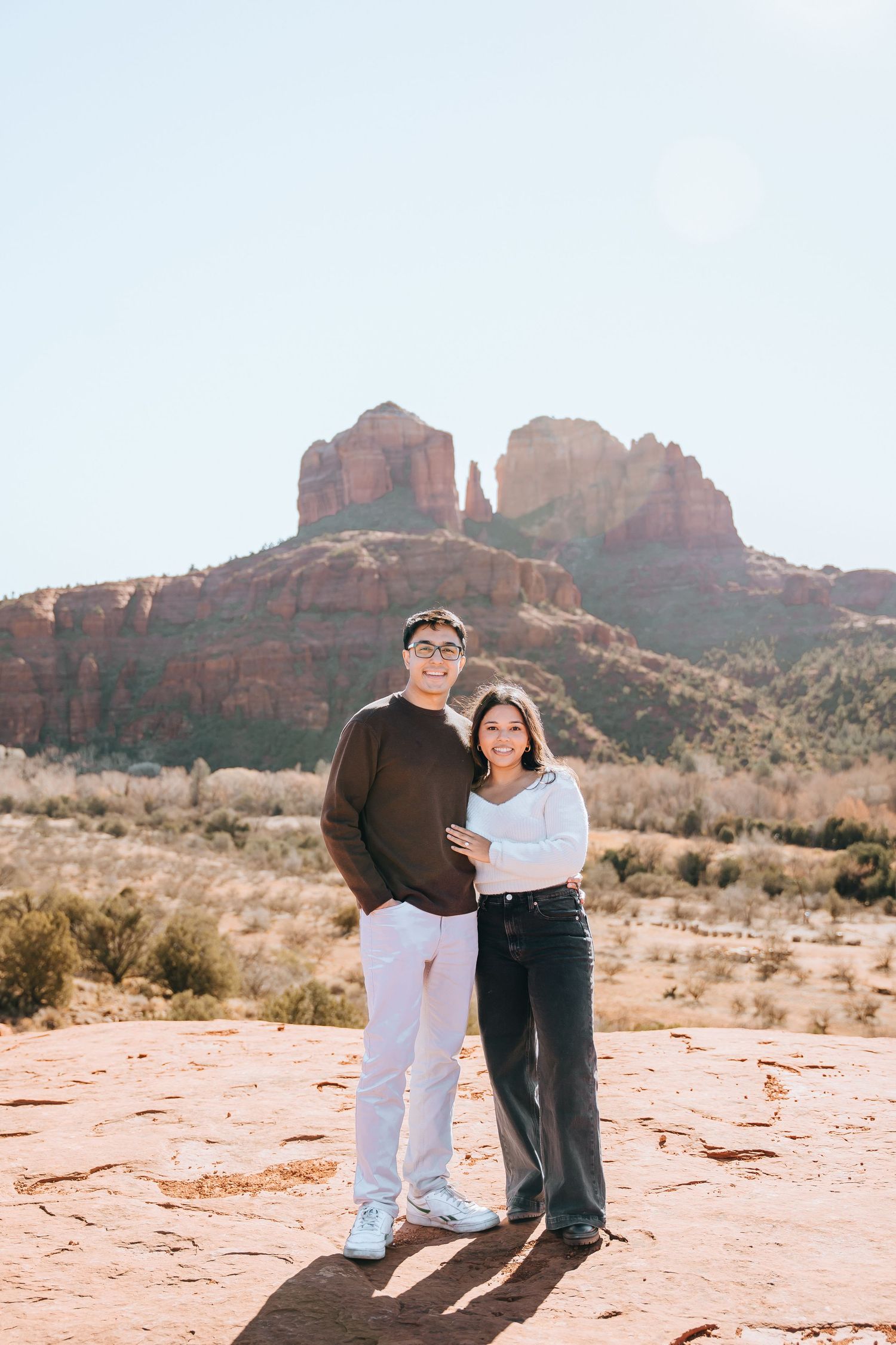Two people embrace while standing on red desert rocks with mountain formations behind them.