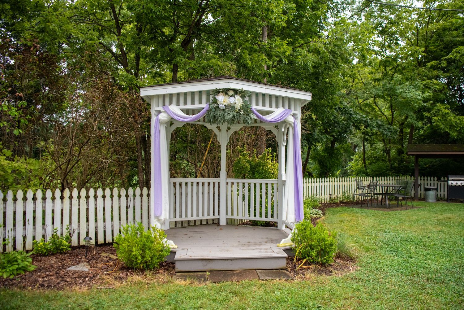 A white wooden gazebo with ornate details stands in a garden setting surrounded by green trees and a white picket fence.