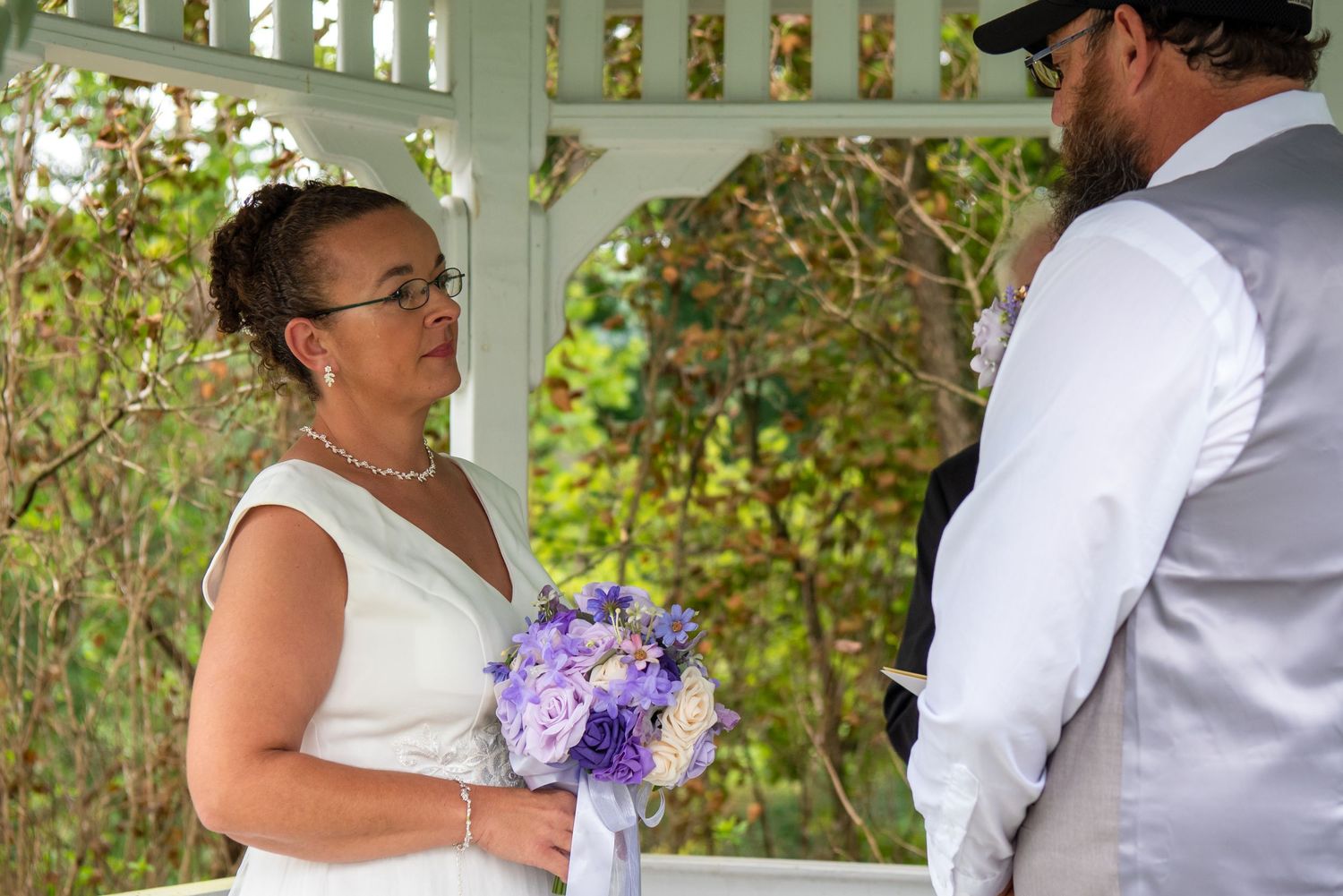 A couple in wedding attire shares a romantic moment under a white pergola surrounded by lush greenery during a ceremony.