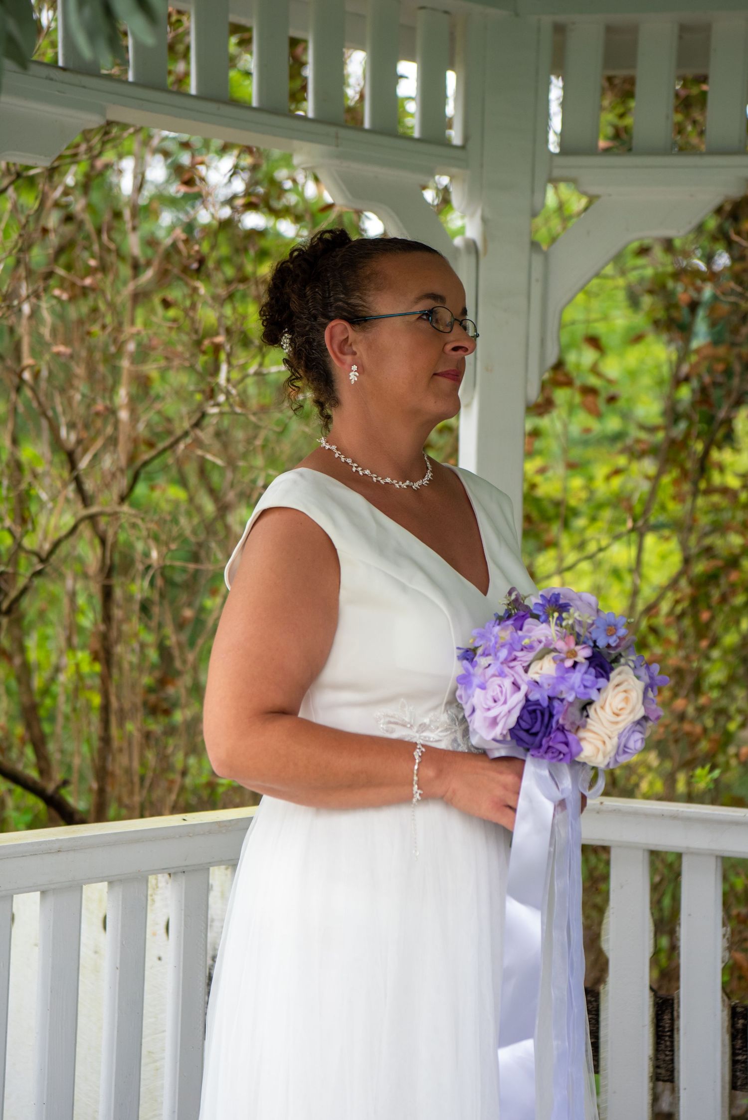 A person in a white wedding dress holds a purple and white floral bouquet while standing in a gazebo.