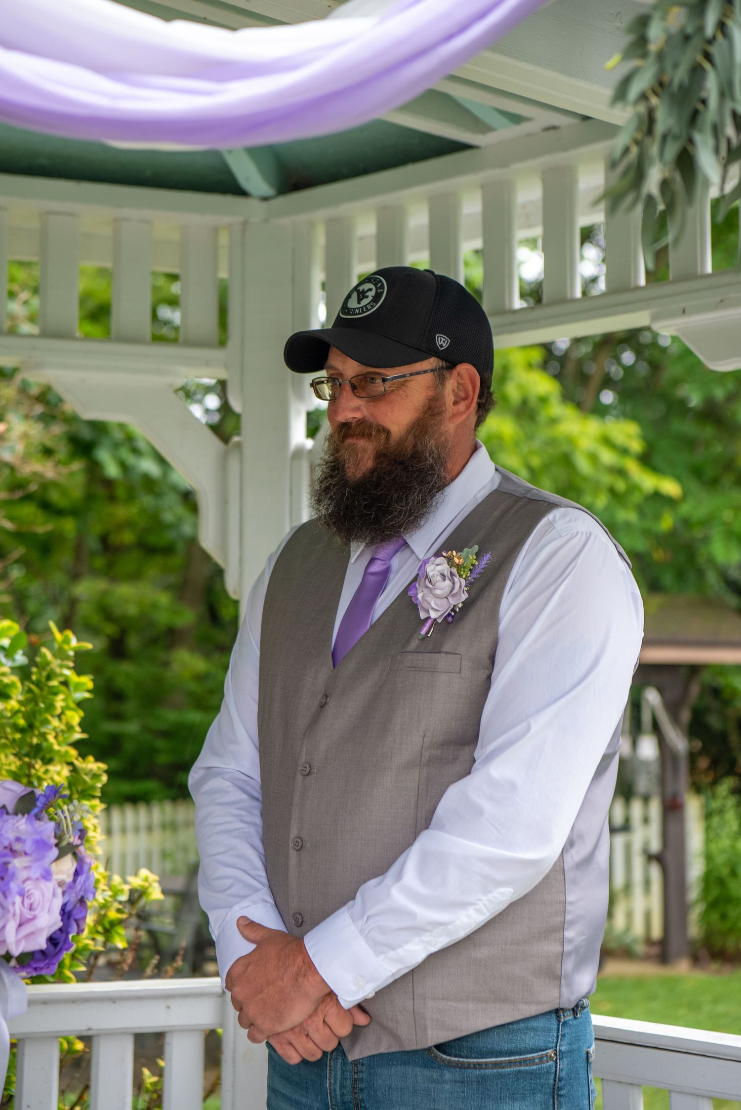 Groom in gray vest and purple tie stands in white gazebo decorated with purple accents for outdoor wedding ceremony.