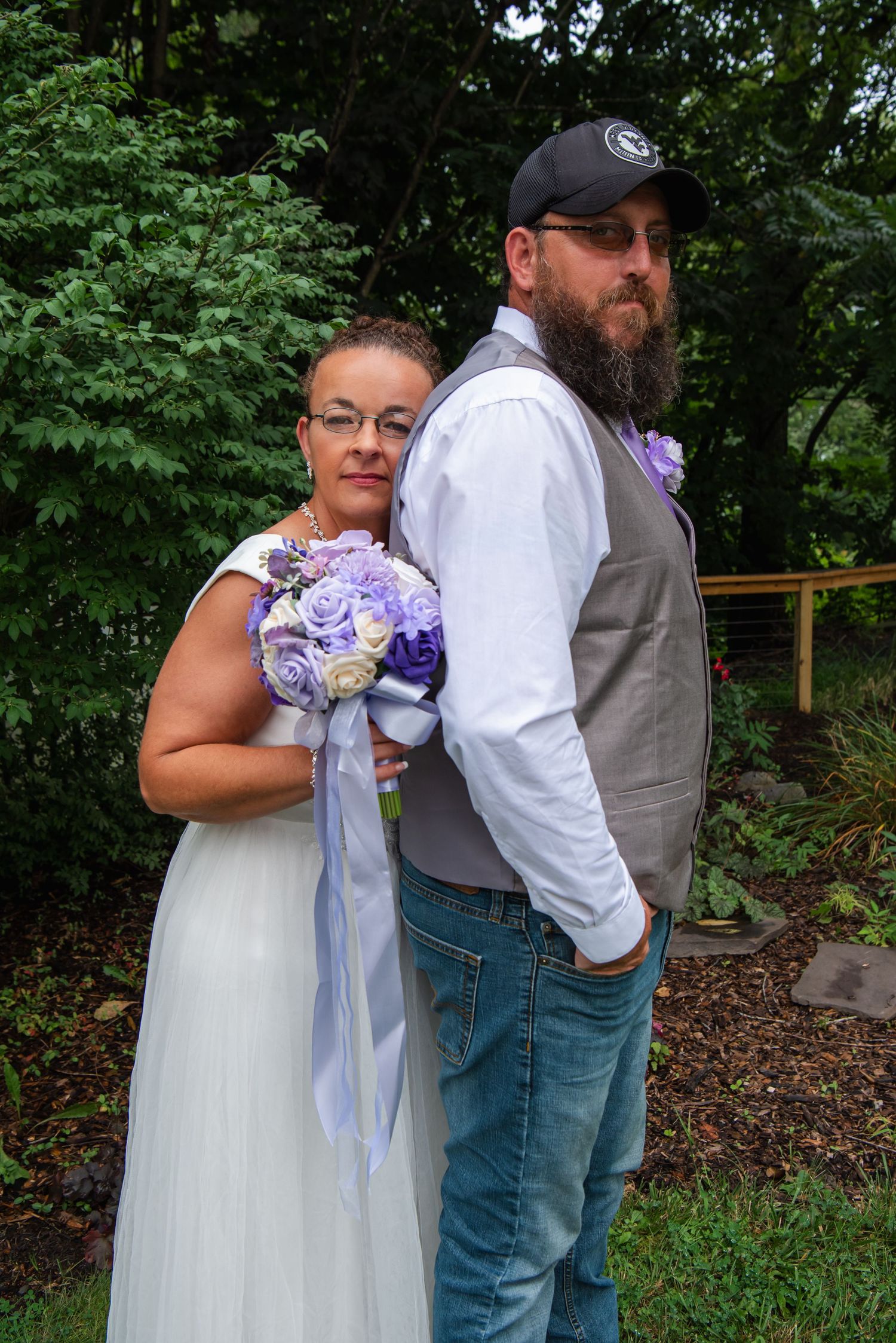 Couple poses back-to-back outdoors at a wedding, with a purple and white bouquet against a green garden background.