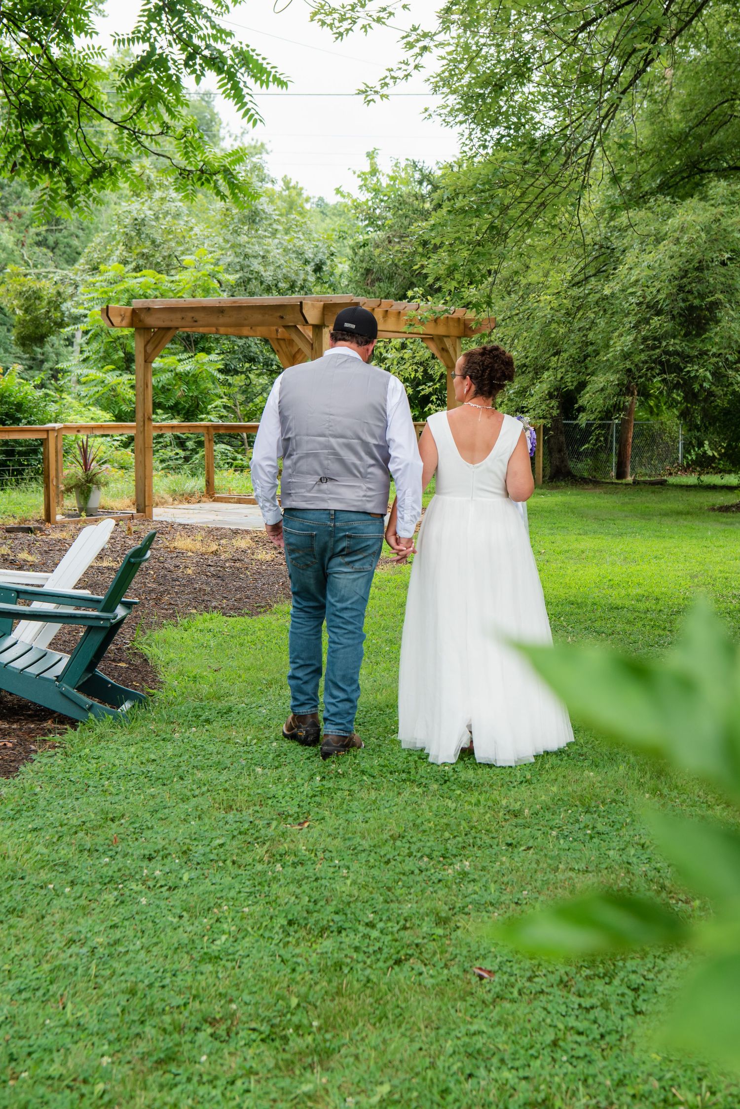 A couple in wedding attire walks through a lush green garden pathway with wooden pergolas and benches.