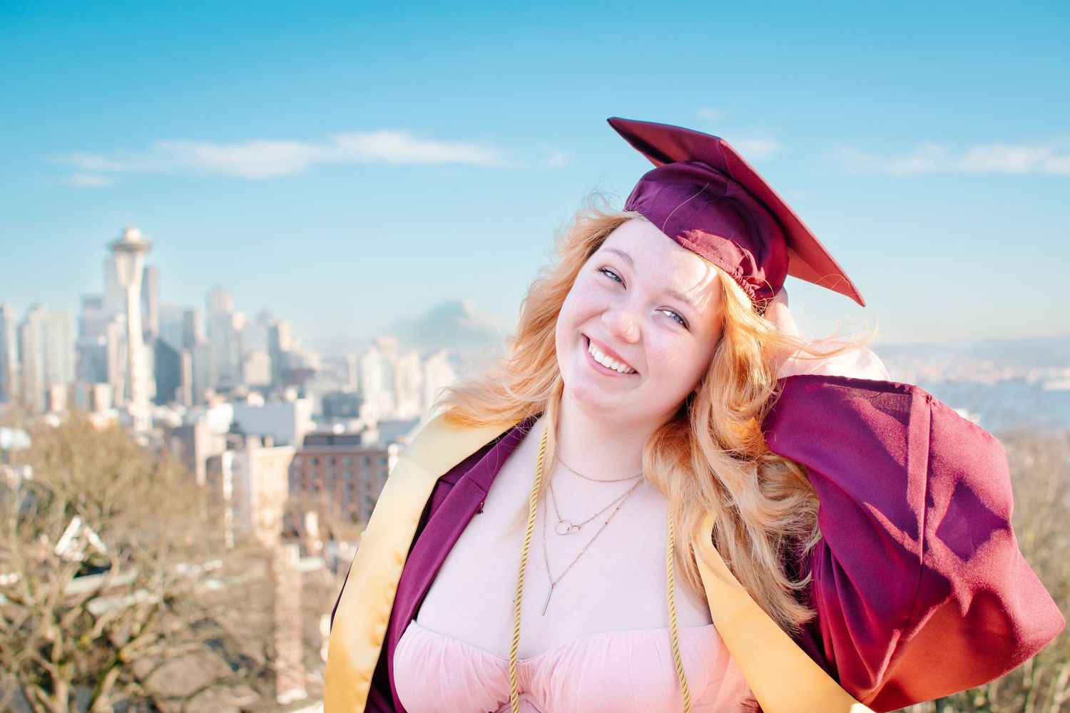 High school graduate in maroon cap and gown posing with Seattle skyline in background