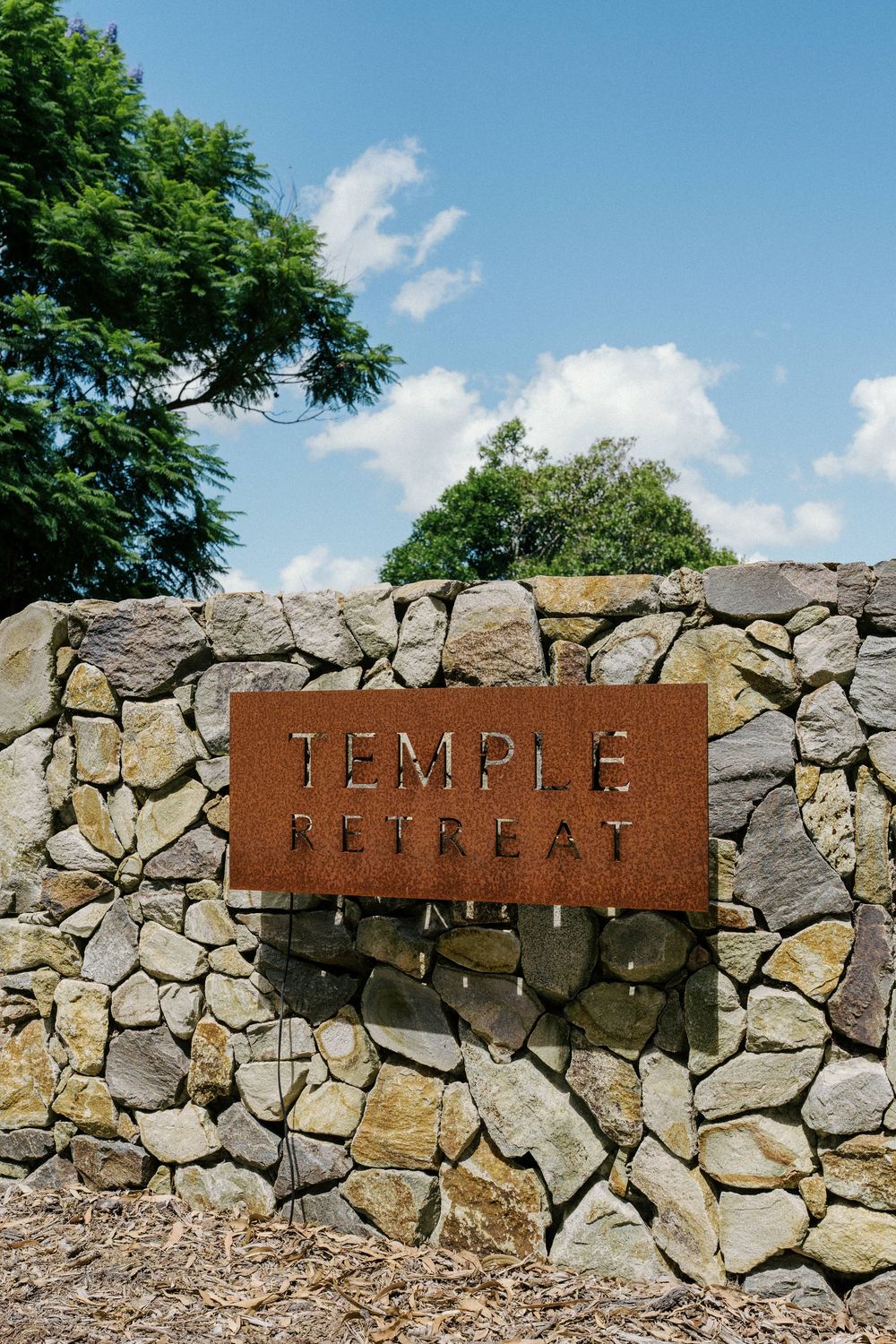 Rustic metal signs spelling out 'TEMPLE RETREAT' mounted on a stone wall against a blue summer sky.