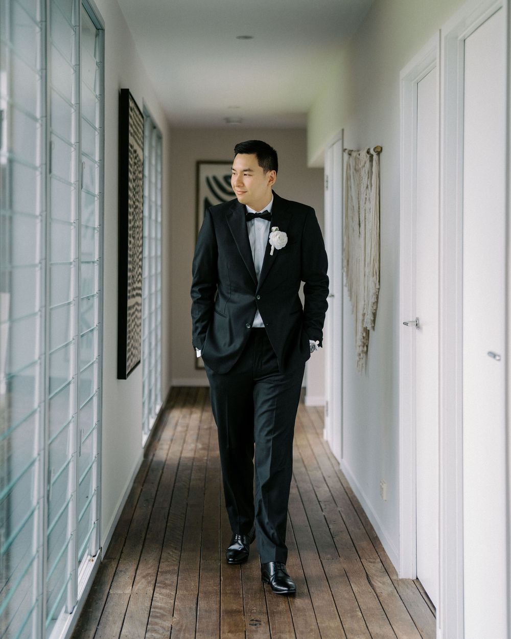 Well-dressed figure in classic black tuxedo and bowtie walks down a bright hallway with wooden floors and white walls.