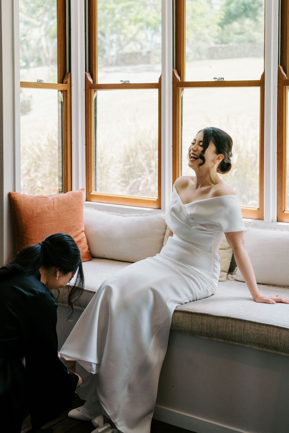 Bride in elegant white off-shoulder dress sitting by window with natural light.