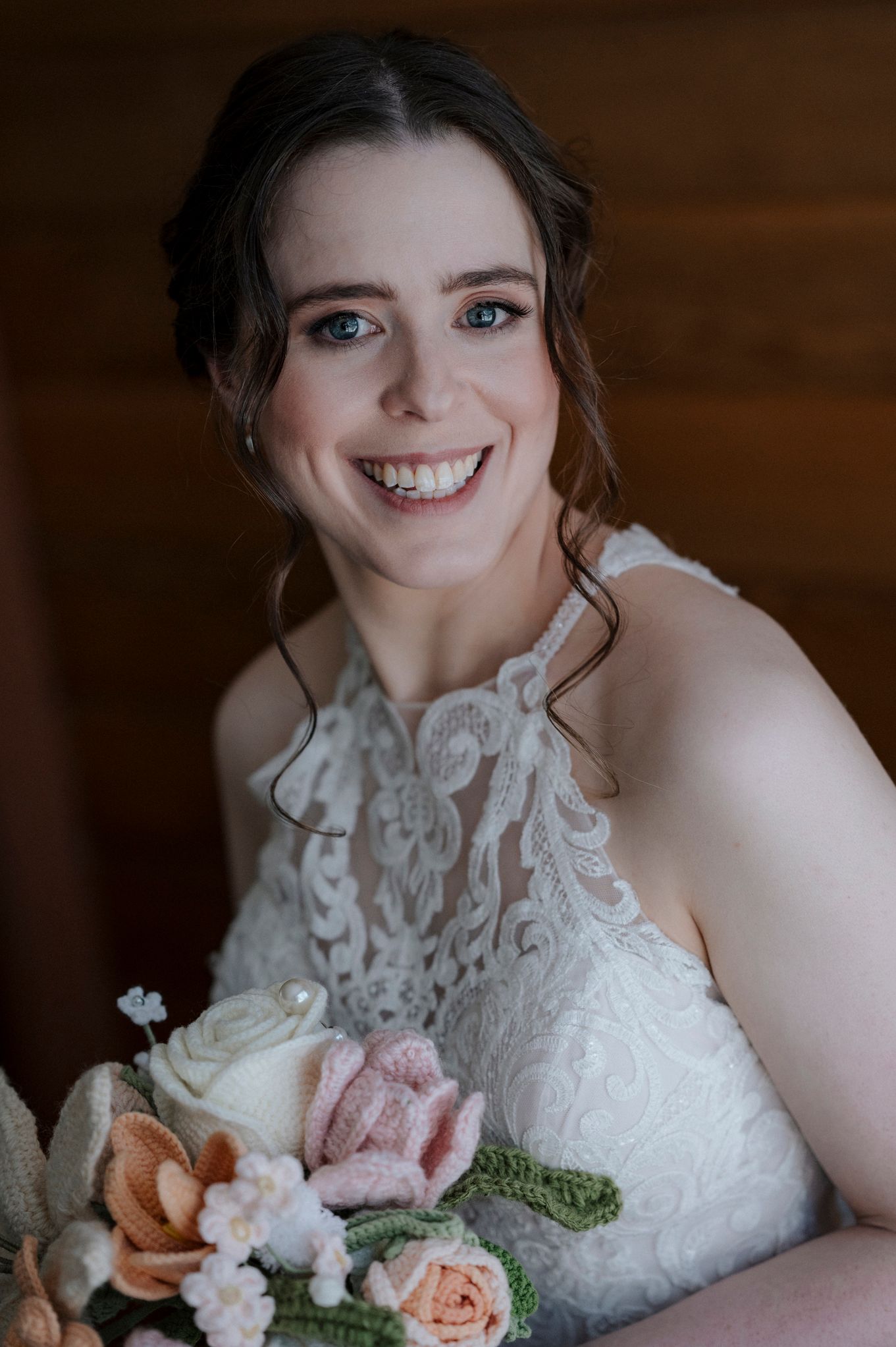 A bride wearing a delicate lace wedding gown smiles while holding a pastel bouquet of roses and flowers.