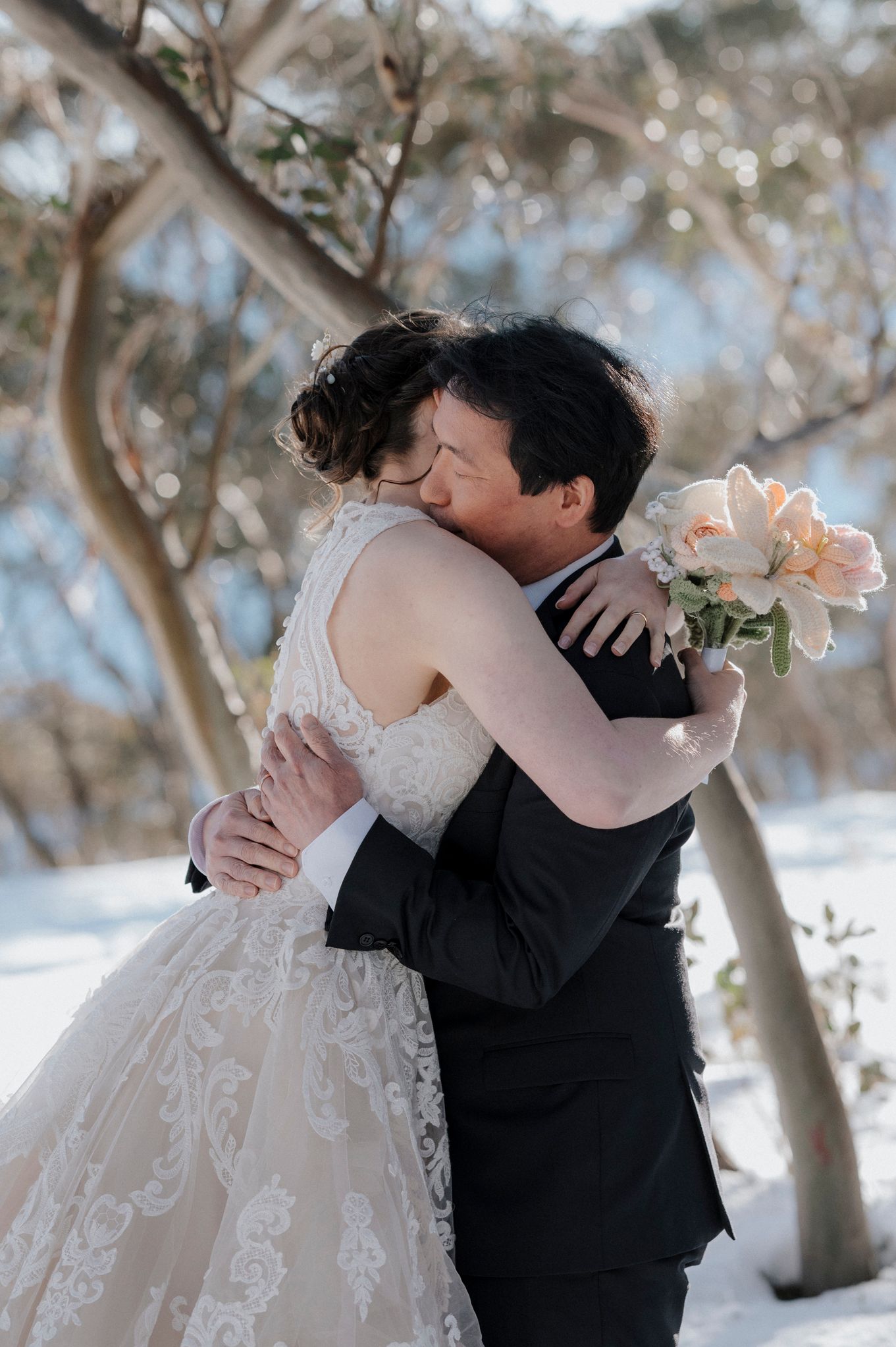 A couple embraces in a tender hug during their winter wedding, with peach roses and snow-covered trees in the background.
