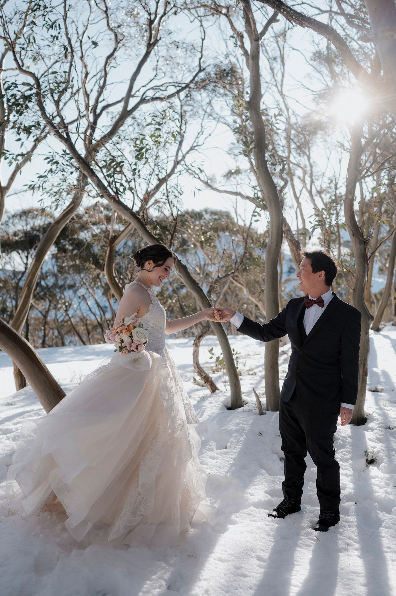 A couple shares playful moments in a snowy winter wonderland beneath snow-covered trees on their wedding day.