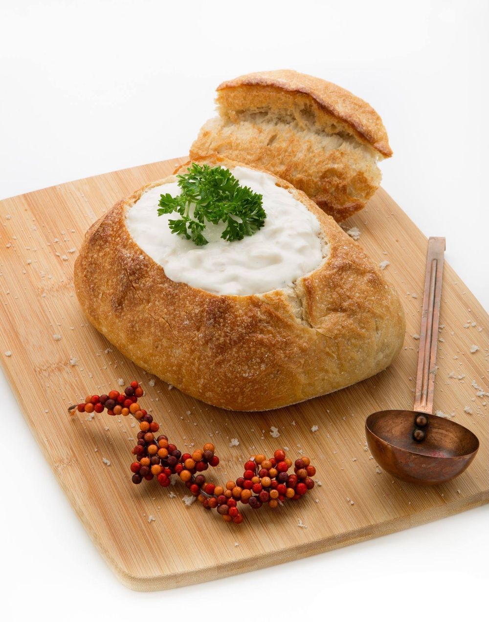 A bread bowl filled with creamy dip garnished with parsley sits on a wooden board with peppercorns and a copper spoon.