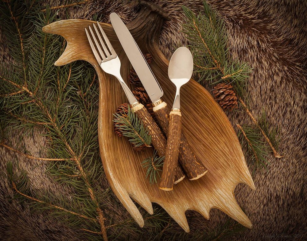 Rustic flatware set arranged on a wooden moose-shaped platter with pine branches and fur accents.