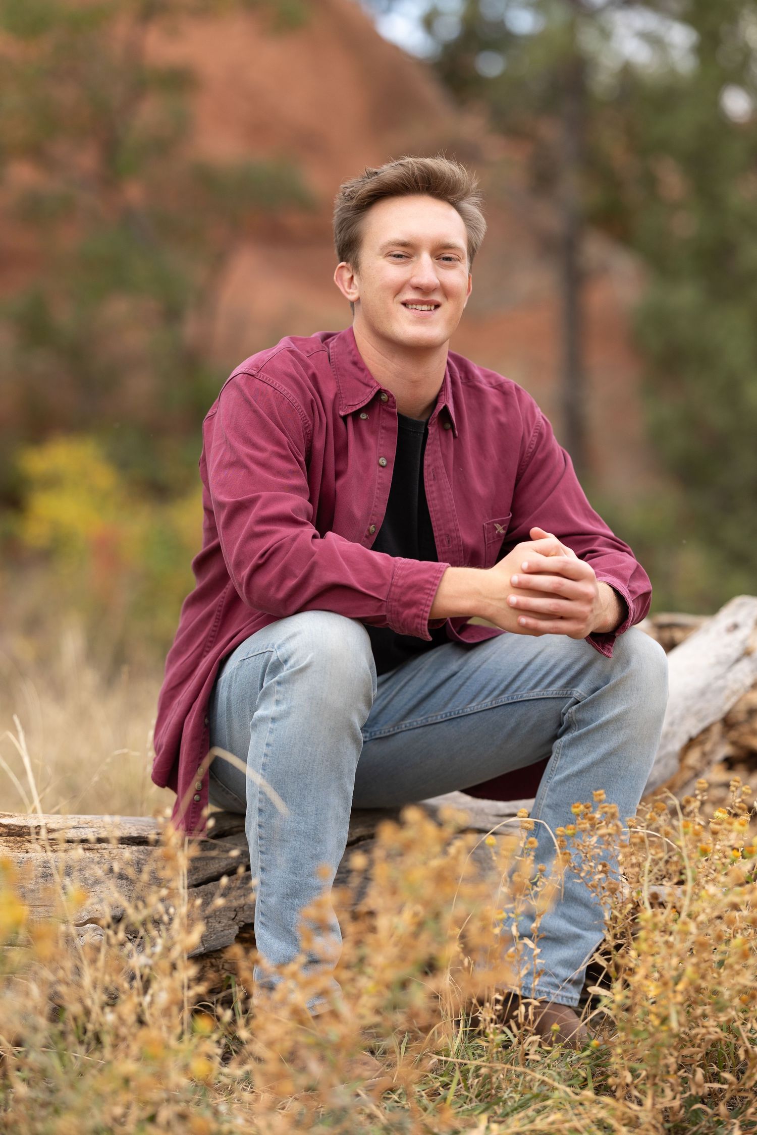 A person in a burgundy jacket and jeans sits casually outdoors among autumn wildflowers and foliage.