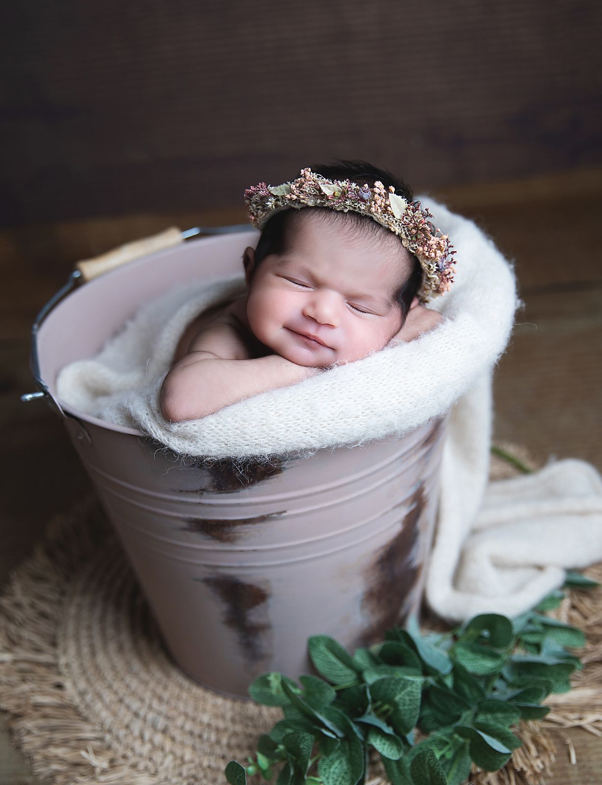 Sleeping newborn during a London newborn session. Nestled in soft white blanket inside pink bucket with green foliage decoration