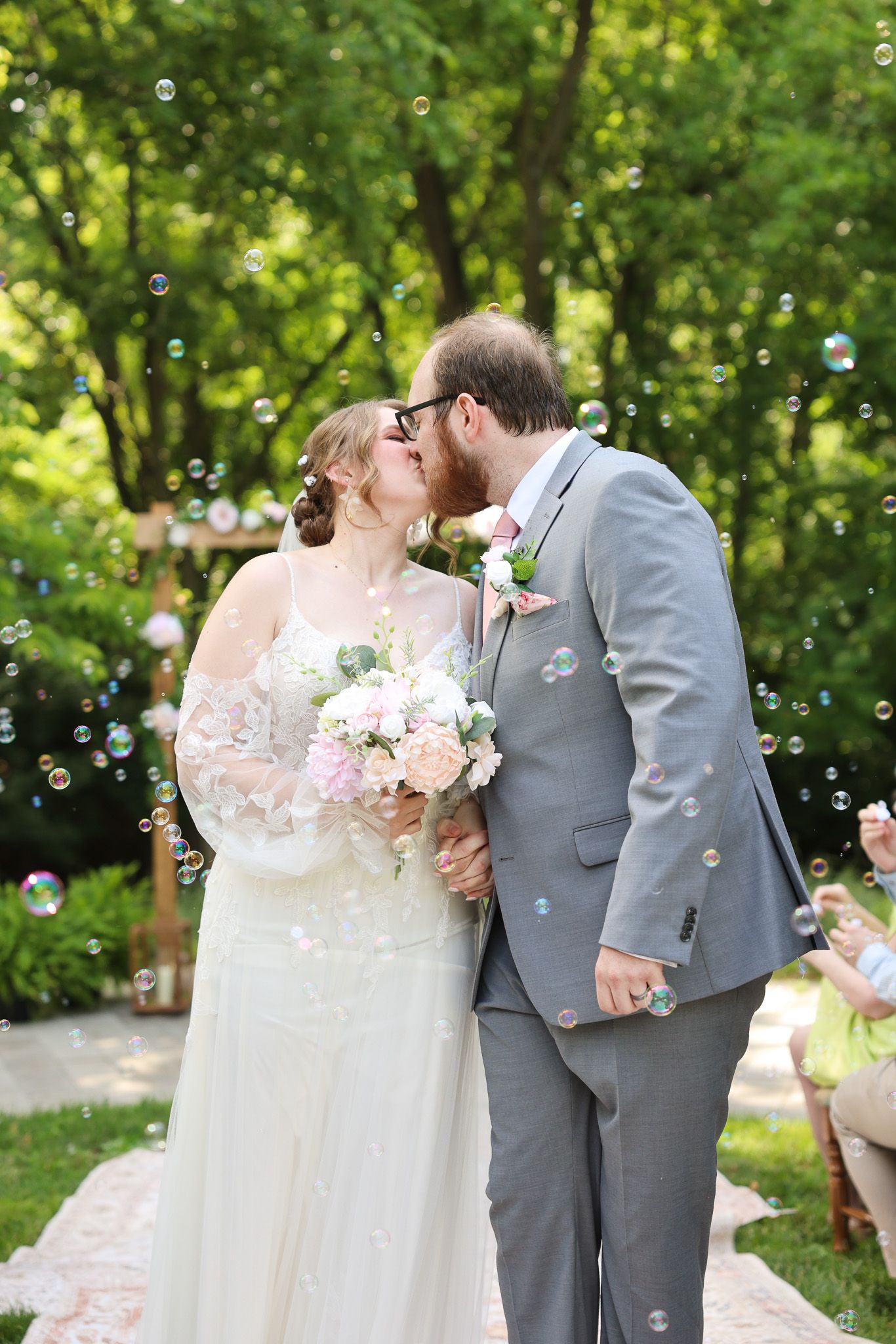 Newlyweds share romantic kiss surrounded by bubbles in lush garden setting.