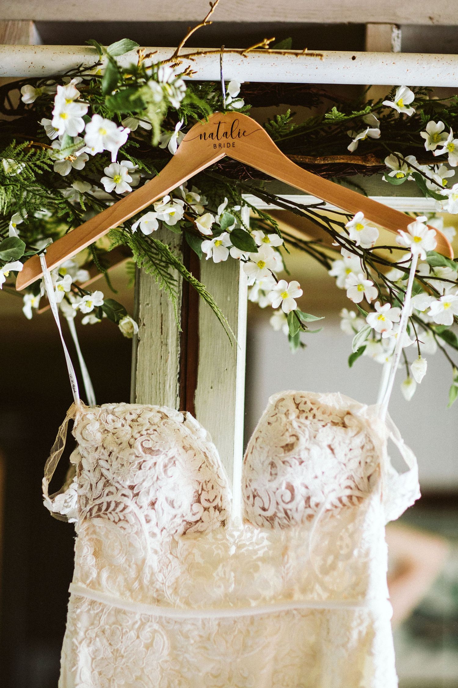 Lace wedding dress hanging on wooden hanger with white floral decorations above.