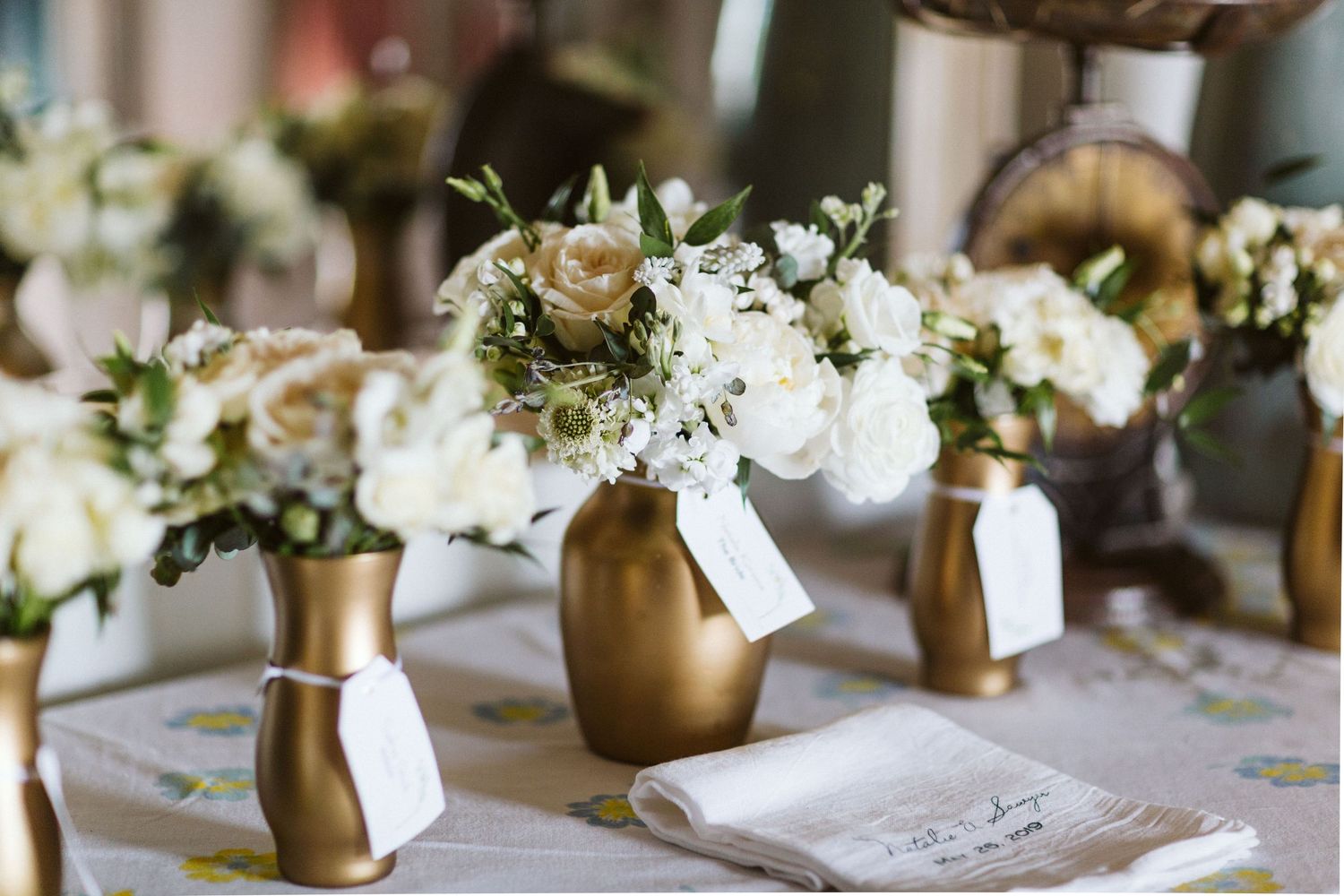 Wedding reception table settings with gold vases and white floral centerpieces.