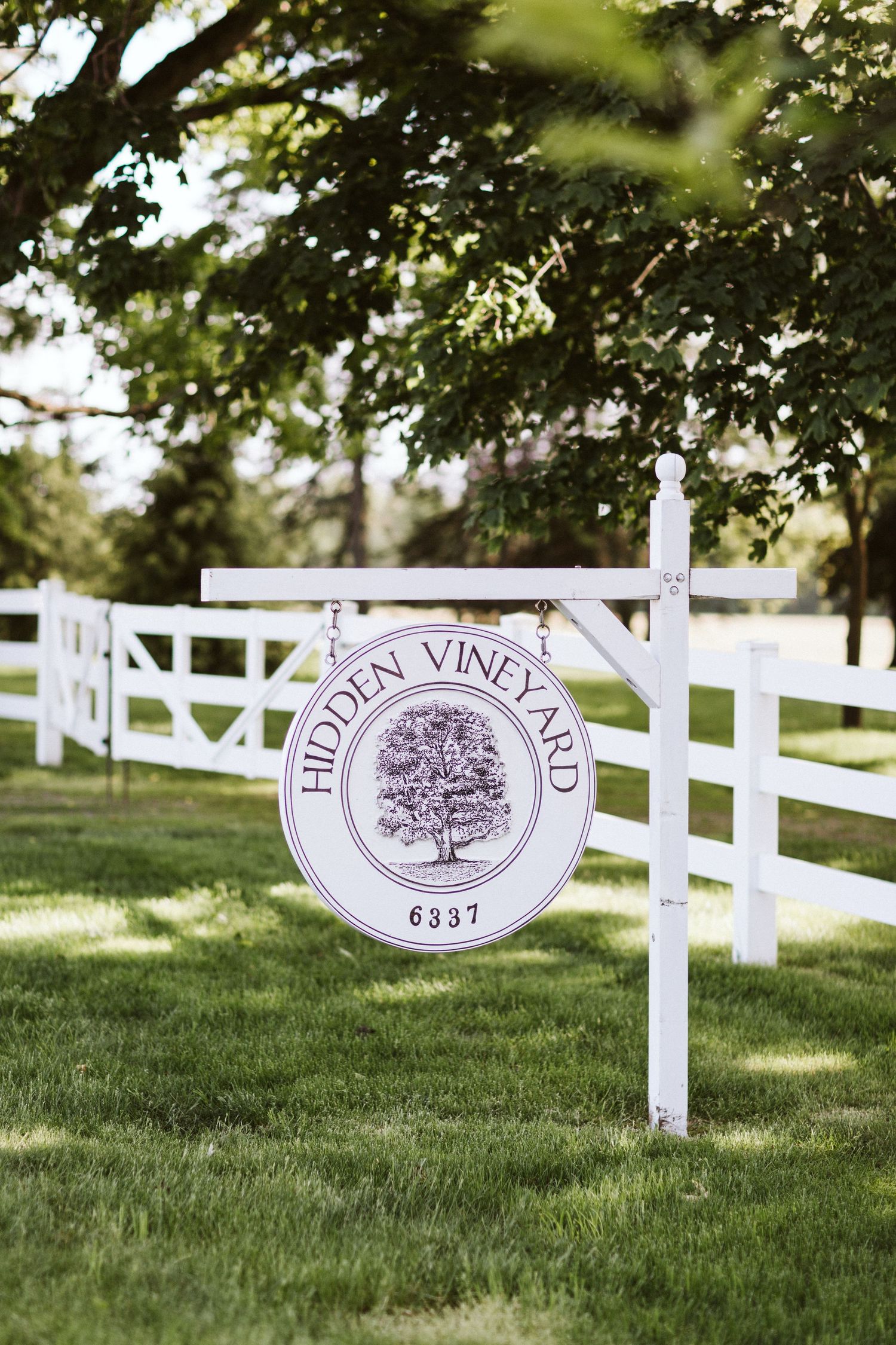 White wooden fence with Hidden Vineyard sign in a lush green garden setting.