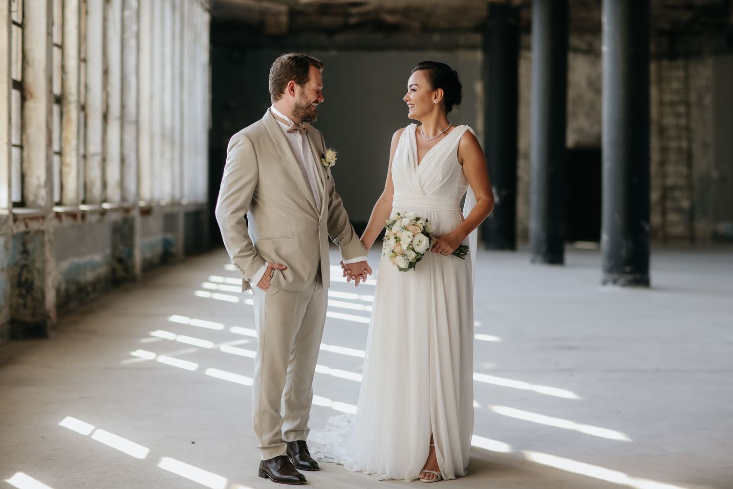 A wedding couple smiles at each other in a sunlit industrial corridor with concrete columns and dramatic shadows.