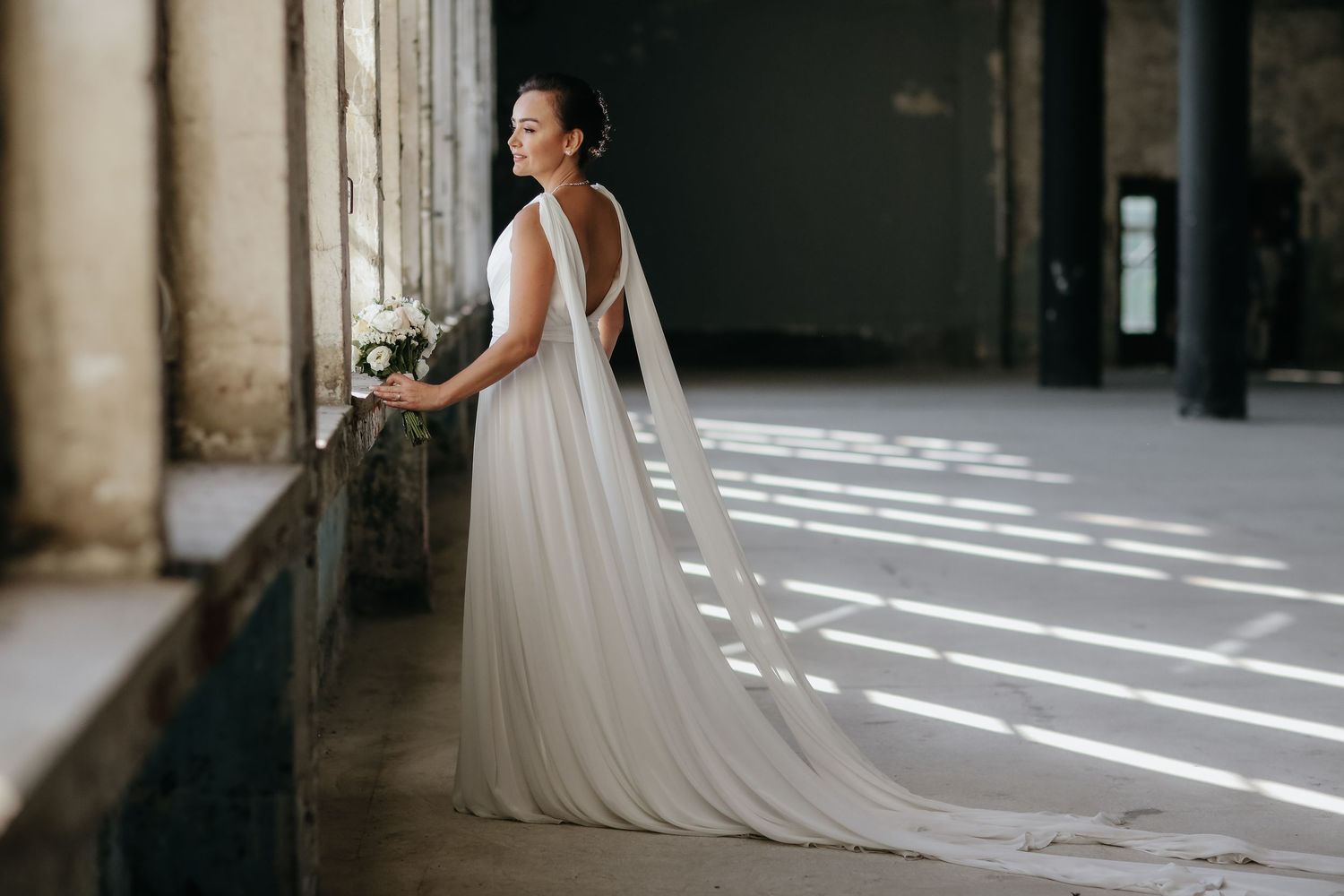 A bride in a flowing white wedding dress stands in dramatic light cast through tall arched windows in an elegant corridor.