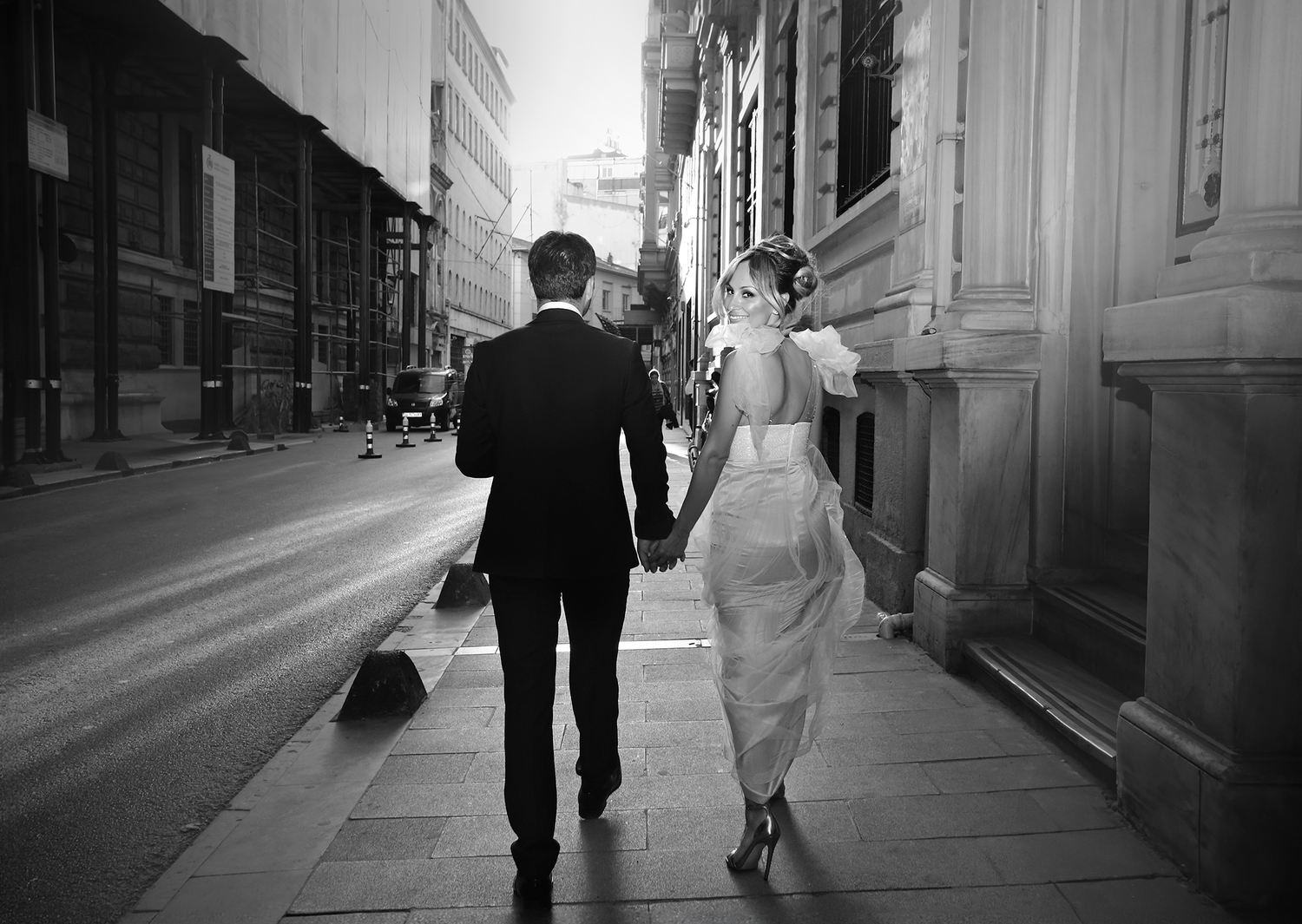 A couple in formal wedding attire walks down a city sidewalk while sharing an intimate moment in black and white.