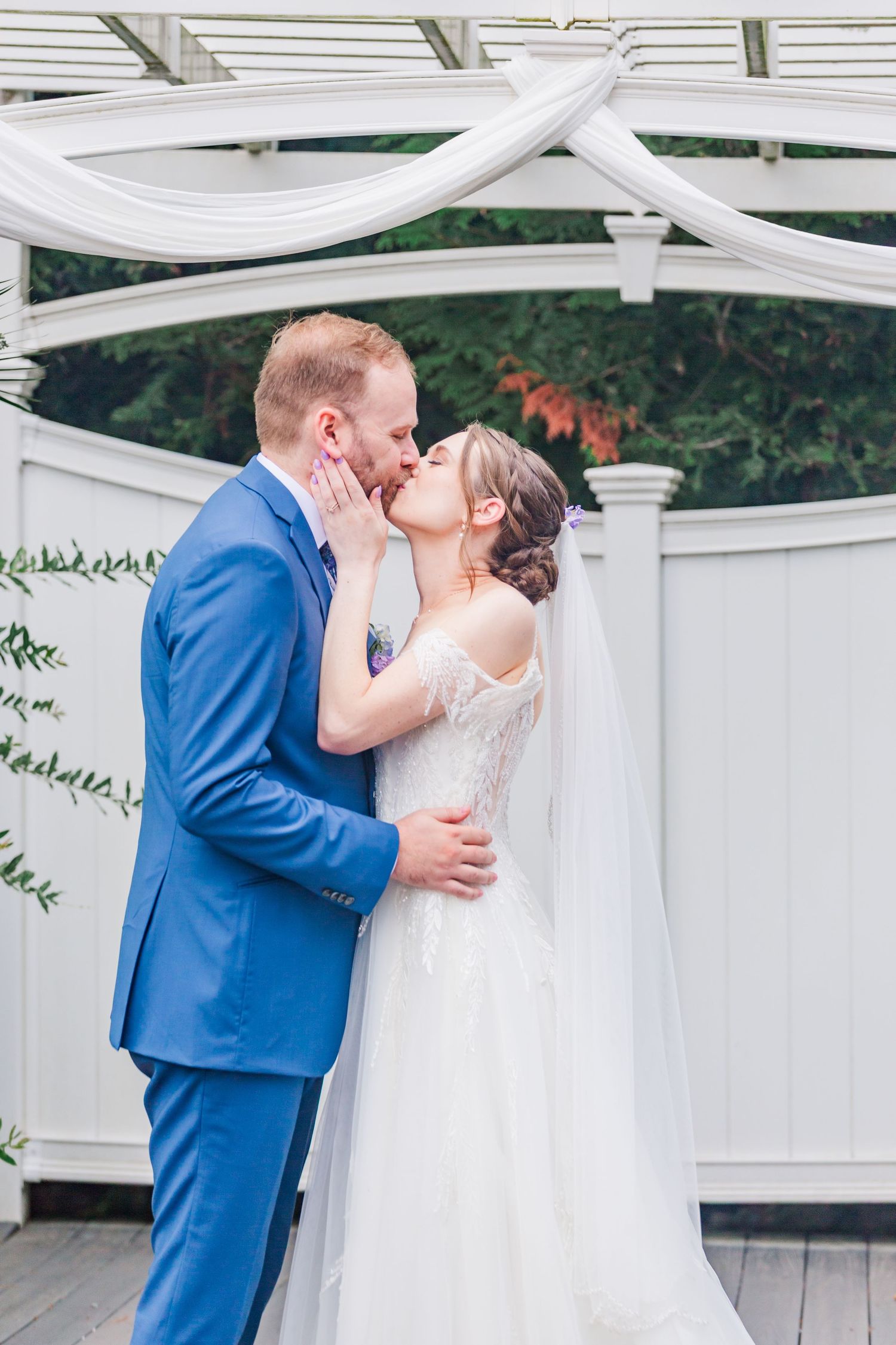 Two people kiss under an elegant white pergola during a wedding ceremony with a bride in white and groom in blue.