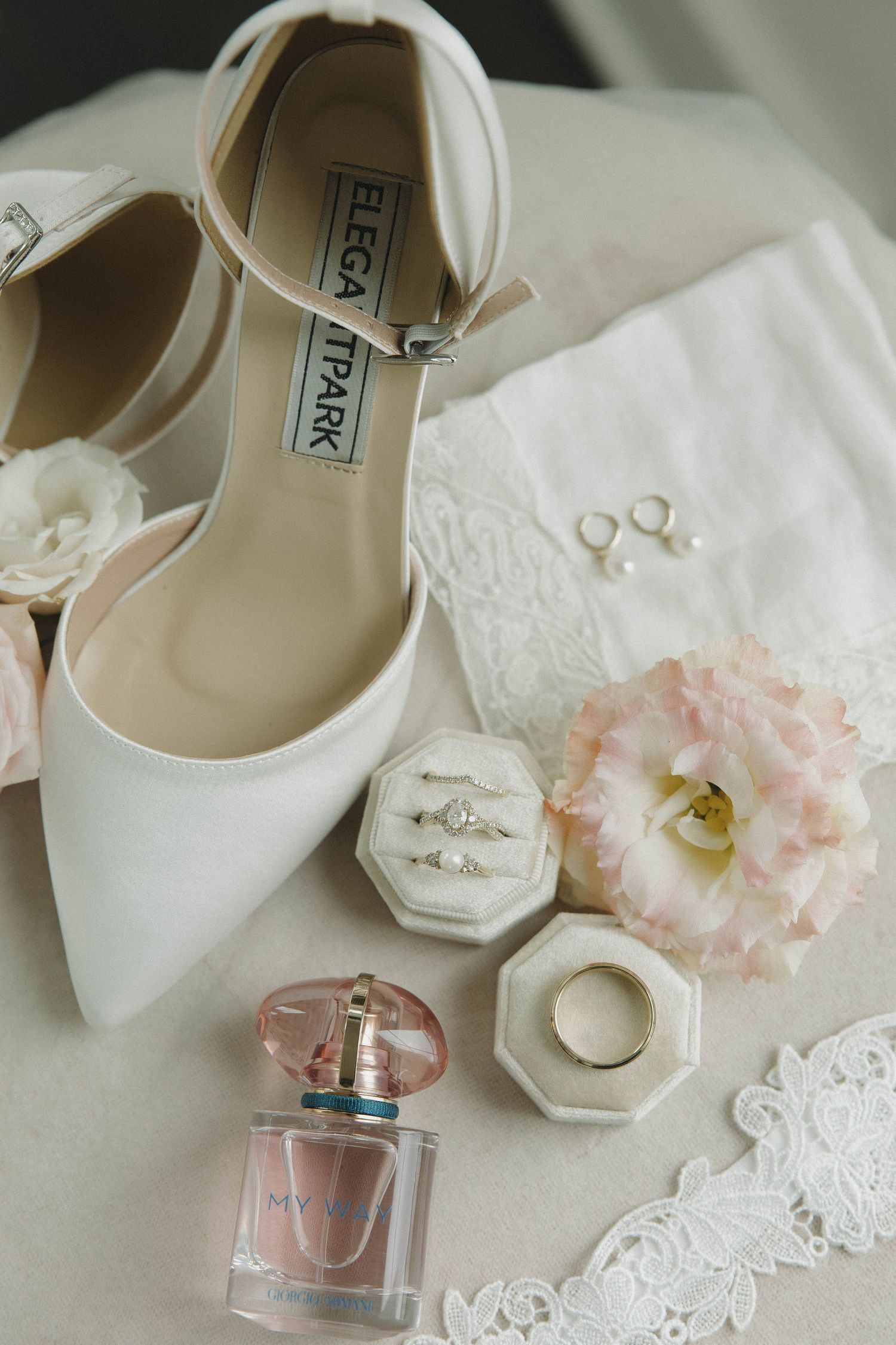 White bridal heels displayed with wedding rings, perfume bottle, and soft pink flowers on lace background.