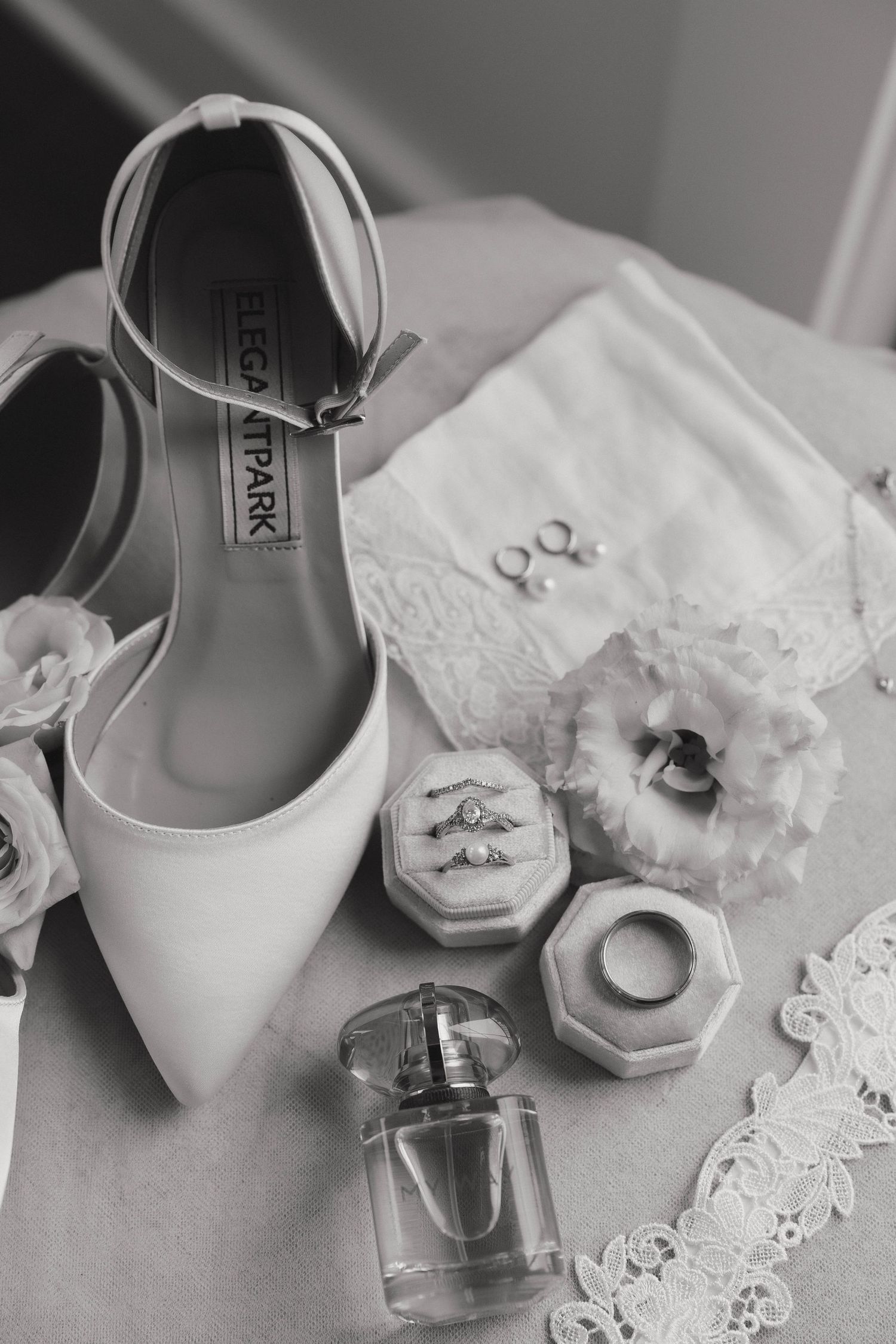Elegant white bridal shoes with perfume bottle, jewelry rings, and lace accents captured in black and white photography.