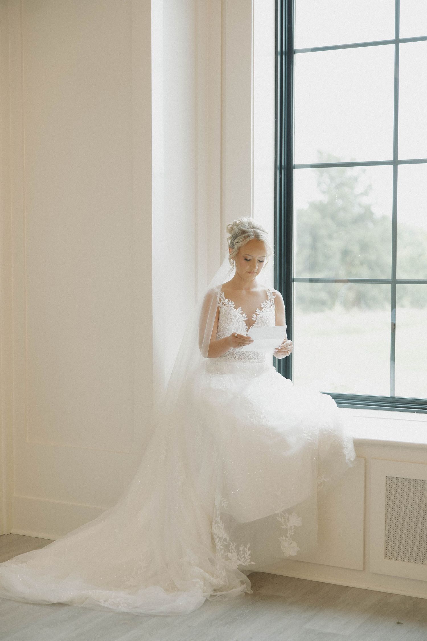 A flowing white wedding dress with intricate lace details is elegantly displayed by large windows in natural light.