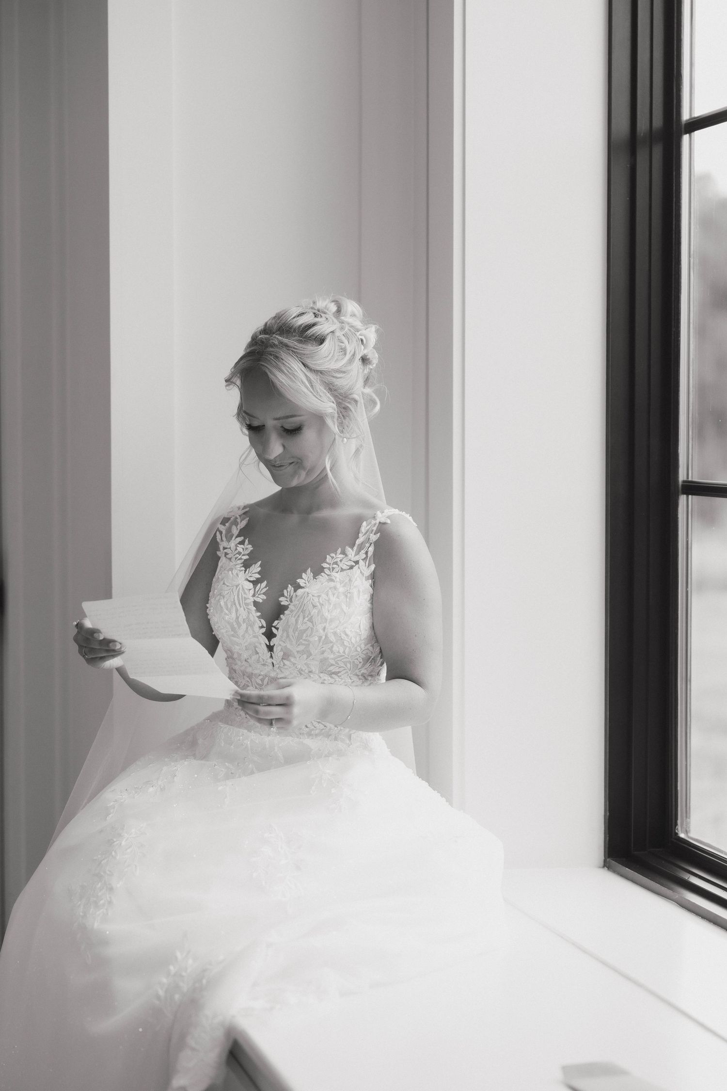 Black and white photo of a bride in a lace wedding gown reading a letter by a window in natural light.