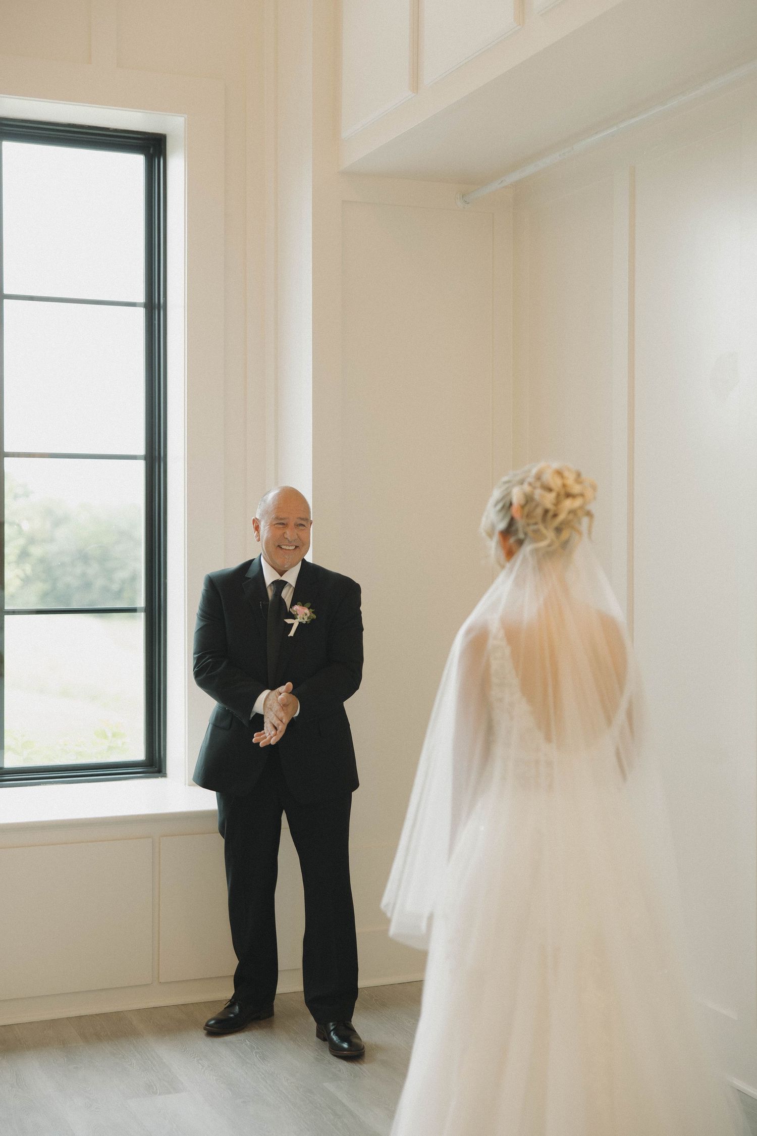 A tender wedding moment captured in a bright room with large windows as a bride in white veil stands with someone in formal attire.