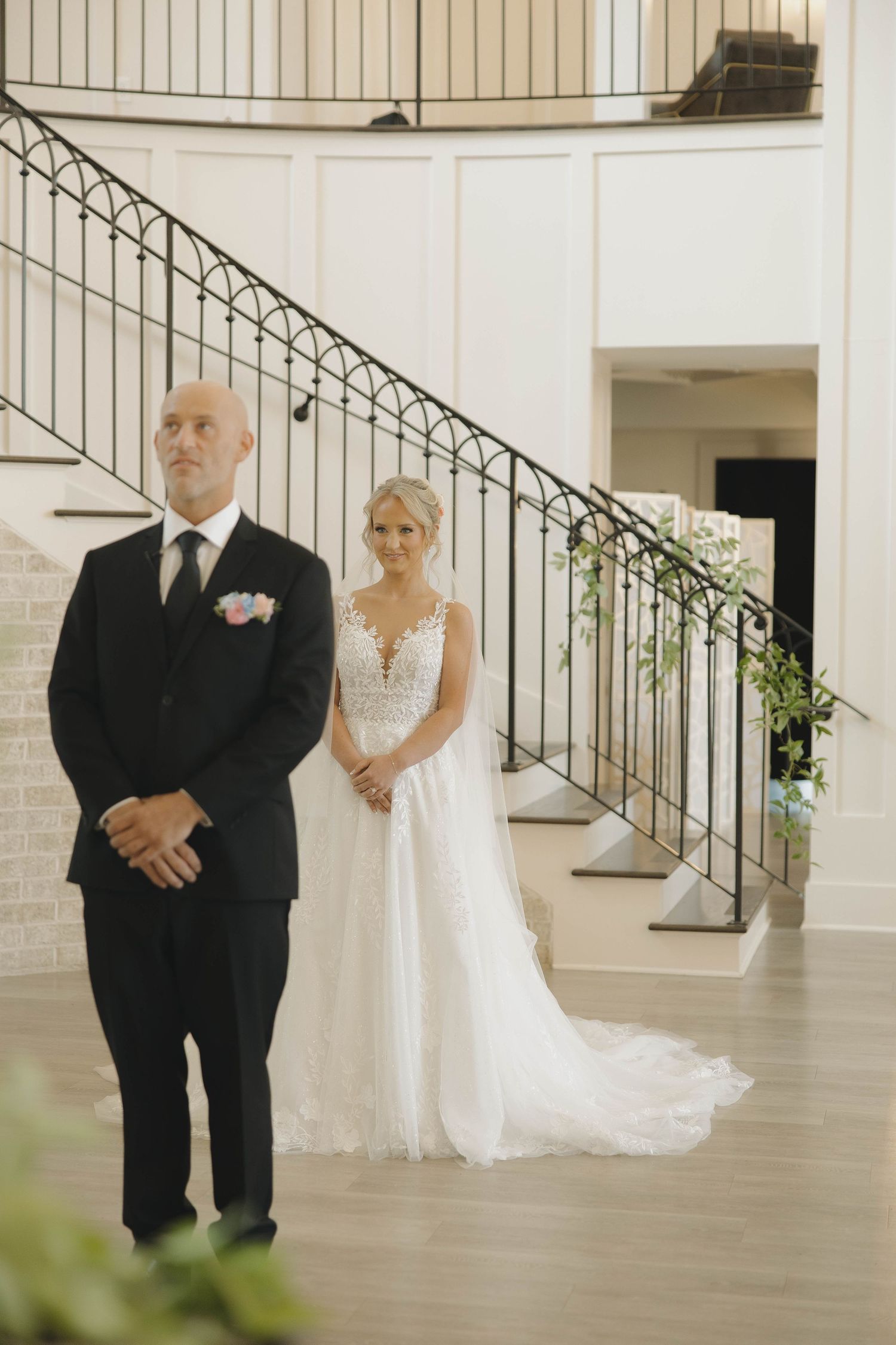 A tender first look moment at a wedding with a black-suited groom and bride in white lace gown by an elegant staircase.