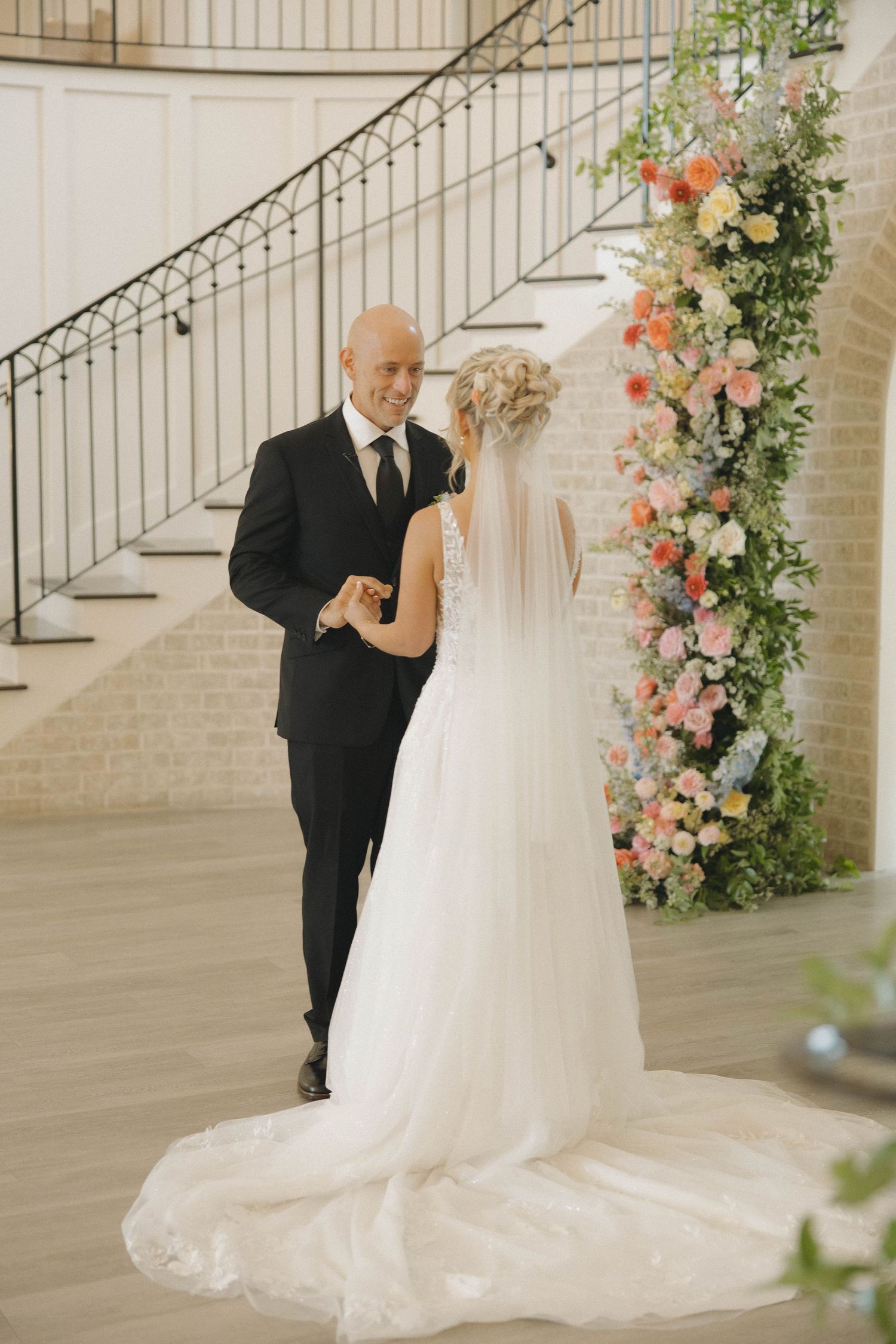 Two people share a tender moment under a floral arch with pink and peach blooms beside an elegant staircase.