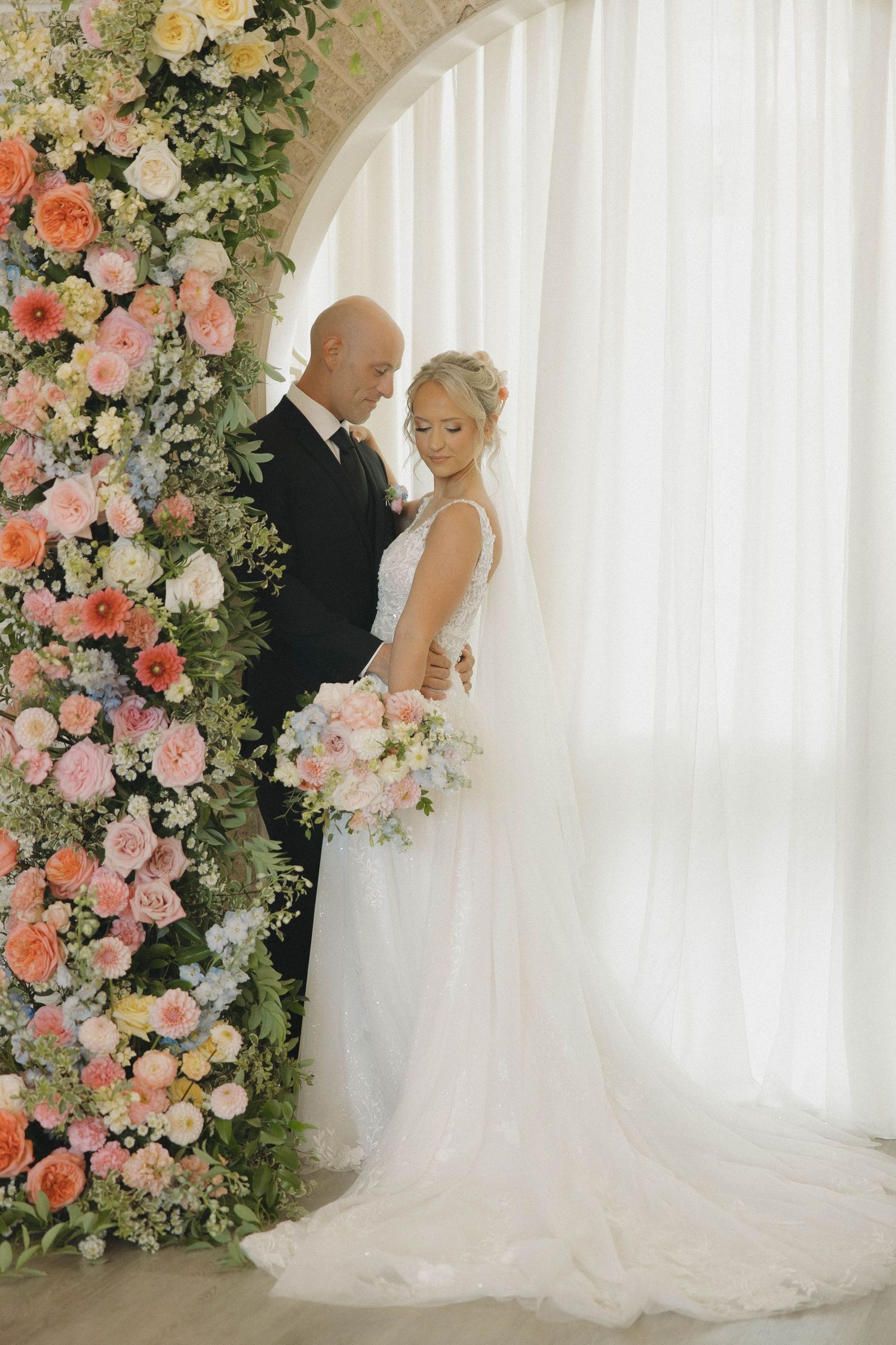 Elegant wedding couple poses under a floral arch with pink and peach roses against a white backdrop.