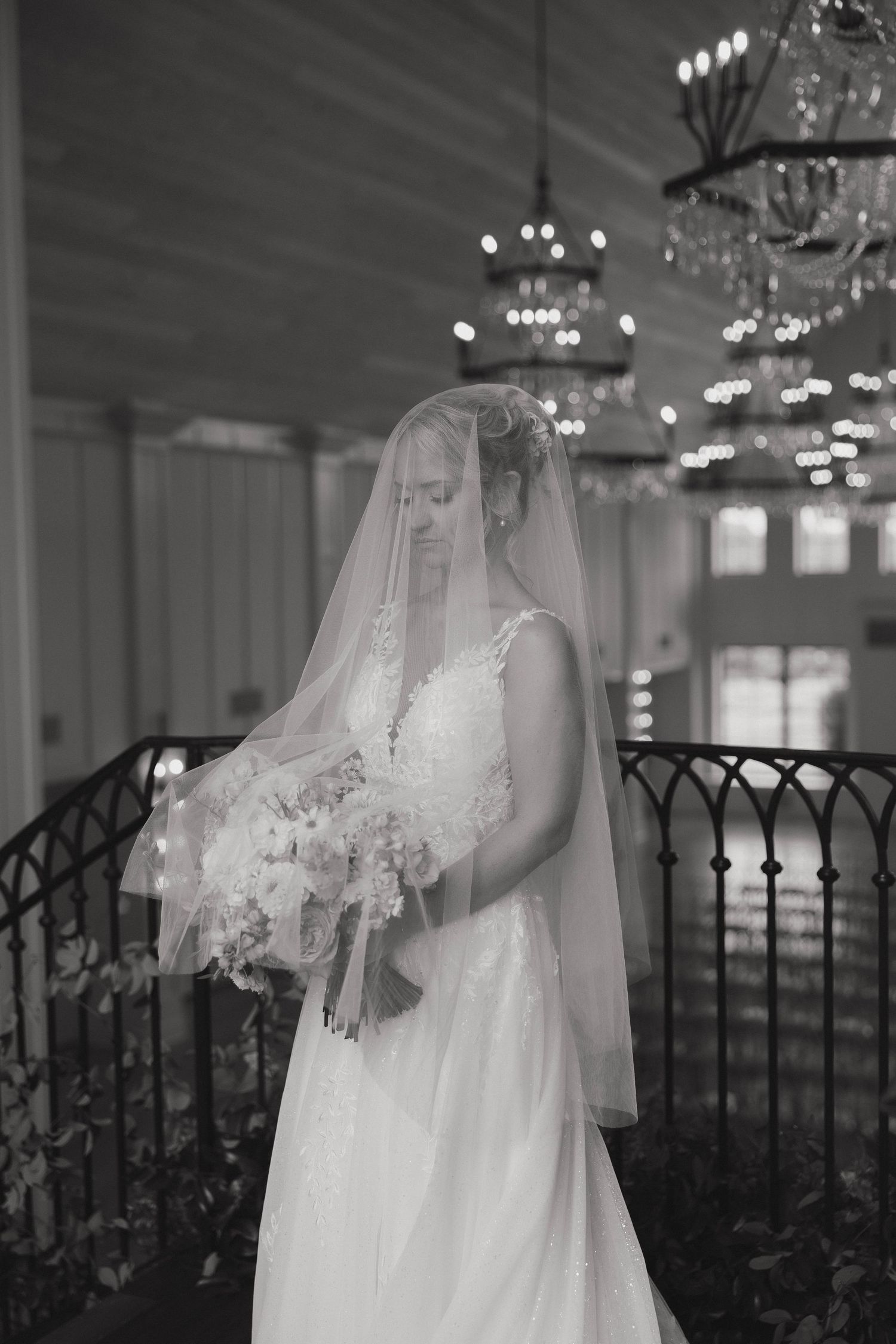 Bride in lace wedding gown holds bouquet beneath crystal chandeliers by decorative iron railing.