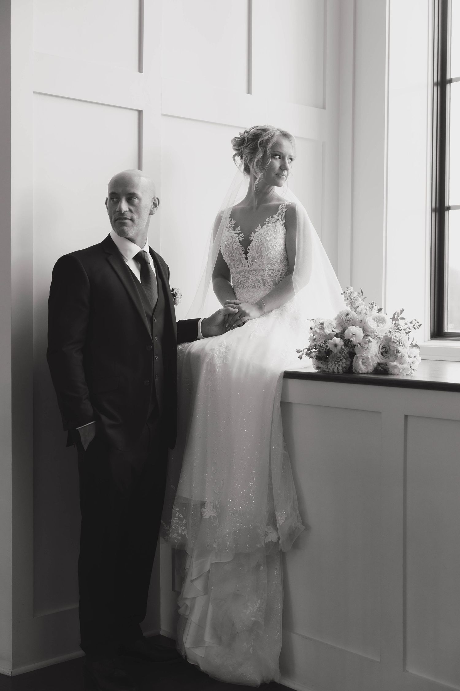 Classic black and white portrait of newlyweds standing by window in formal wedding attire.