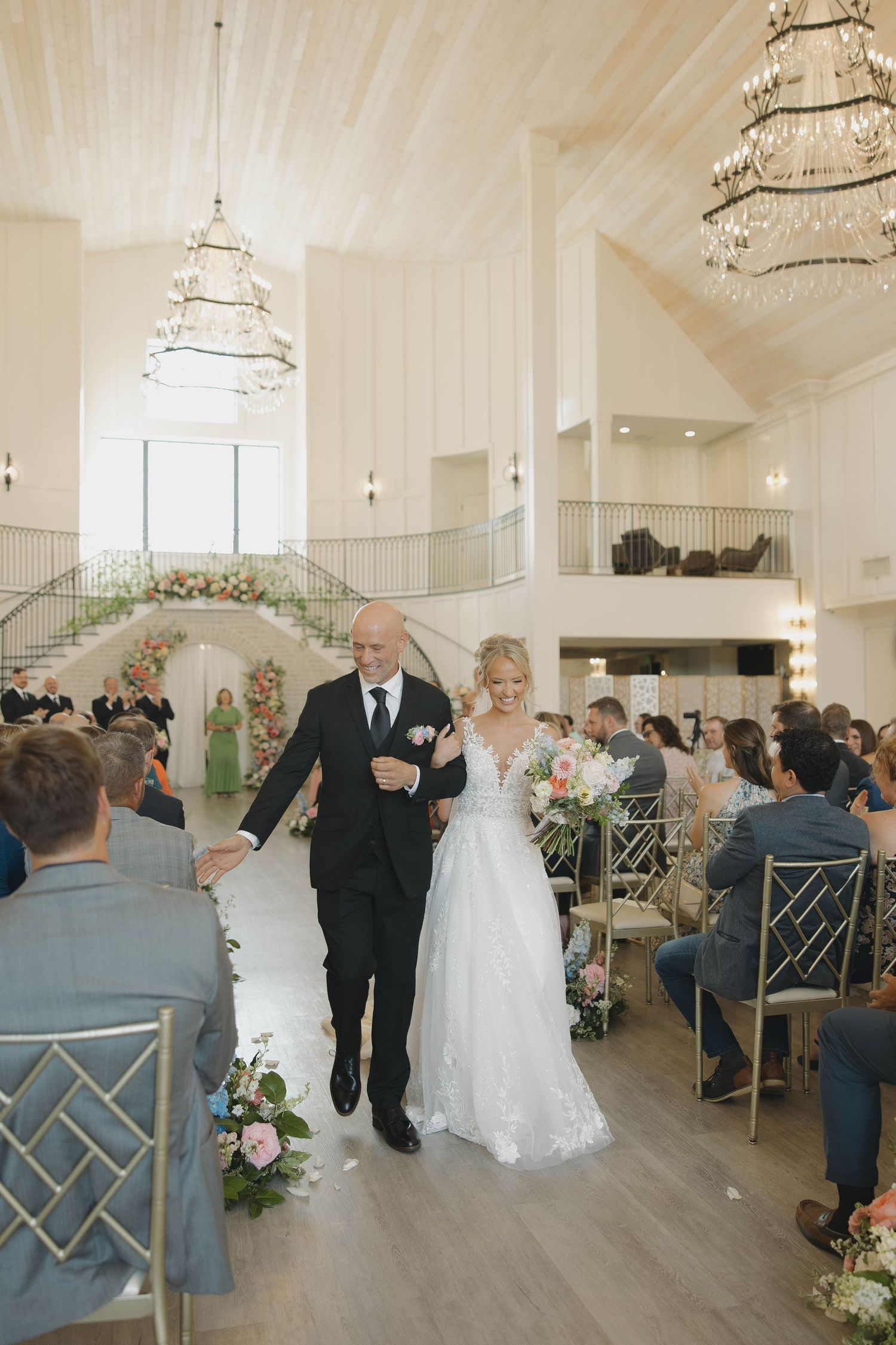 A wedding ceremony taking place in an elegant white venue with crystal chandeliers and light wooden floors.