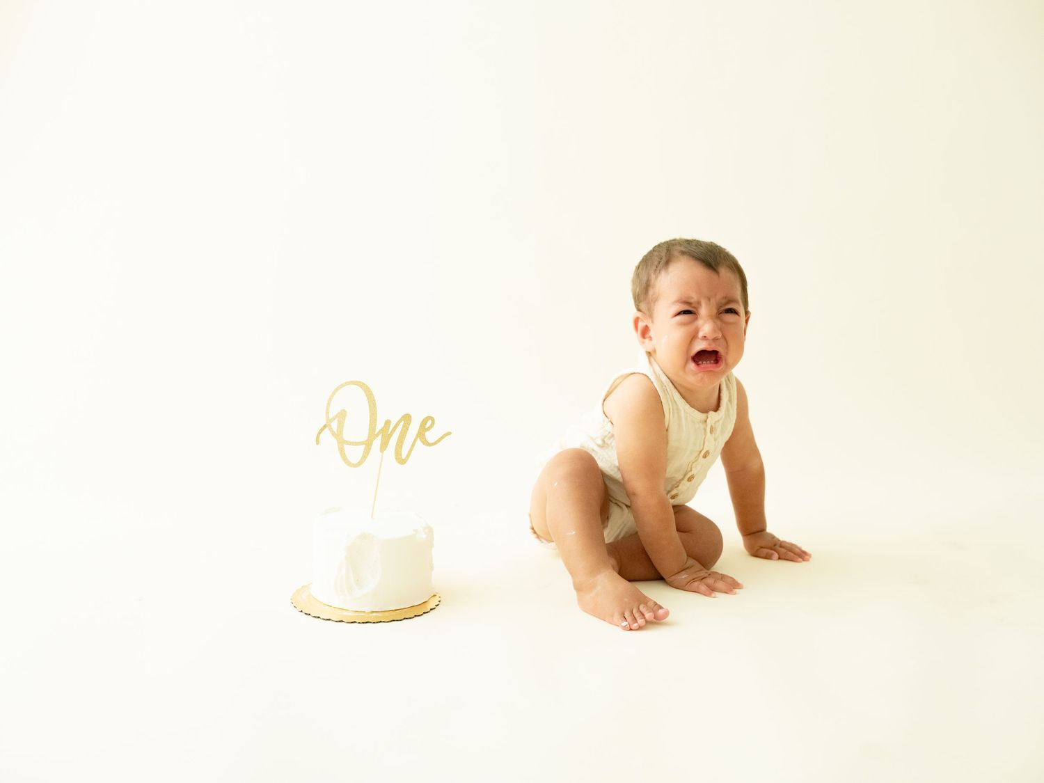 Baby in white onesie lies on light background next to first birthday cake in neutral color palette.