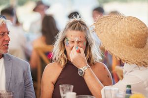 People gather at a sunlit outdoor event, laughing and socializing while holding drinks and wearing summer attire.