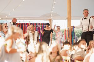 Wedding guests celebrate and dance together under a white tent with suspended lights and clothing racks visible in background.