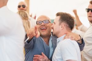 Two people share a joyful embrace while dancing at an outdoor party with other guests visible in sunglasses.