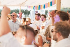 Wedding guests celebrate and laugh together under a colorful banner at an outdoor reception tent.