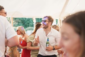 Party guests mingle and socialize under a white tent at an outdoor summer celebration with drinks in hand.