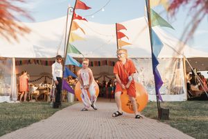 Playful festival scene with colorful flags and a large orange bounce ball against white tents on a grassy field.